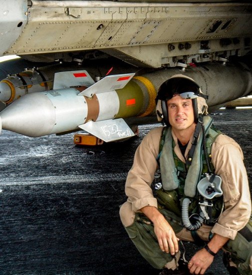 A man in military or pilot gear crouches in front of a military aircraft with a missile attached underneath.