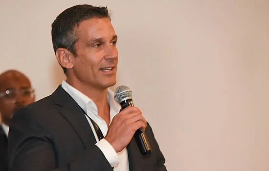 A man in a dark suit and white shirt speaking into a microphone at an indoor event.
