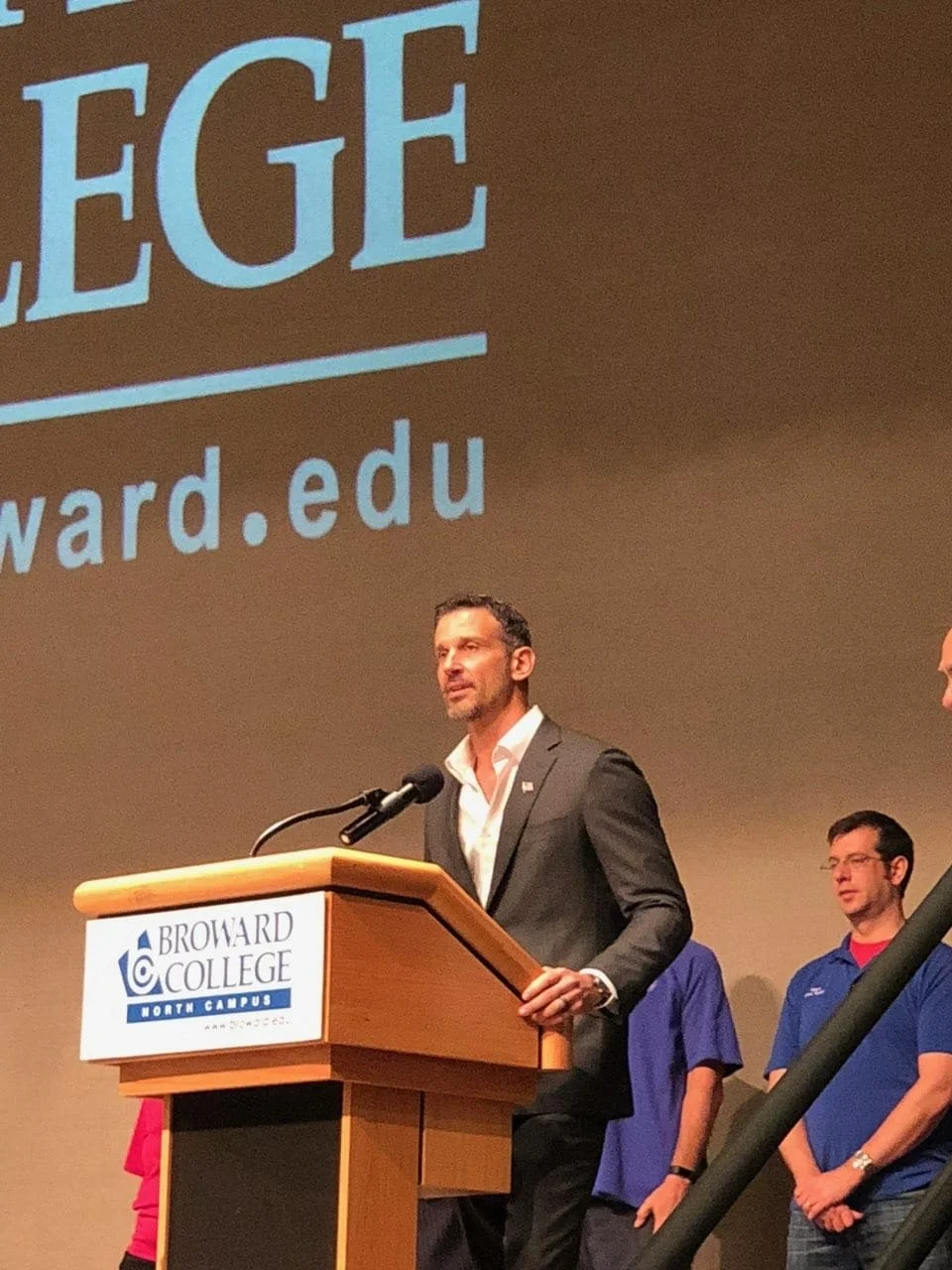 A man in a dark suit and white shirt is speaking at a podium with a microphone at Broward College North Campus. Behind him, there are people in casual blue and purple shirts standing on stage. A large projection screen displays part of the word 'LEADING' and the website 'broward.edu'.