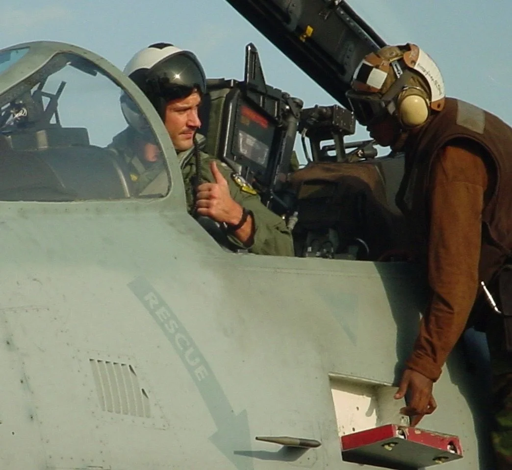 A pilot inside a fighter jet cockpit talking to a crew member outside, who is pointing at a compartment on the aircraft's exterior.