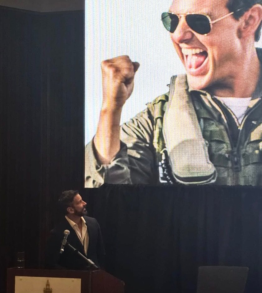 A man stands at a podium with a microphone, looking up at a large screen displaying a photo of a smiling person wearing sunglasses and a military-style jacket, making a fist gesture.