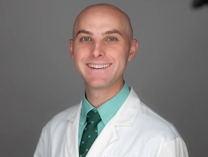Portrait of a smiling male doctor wearing a white coat, green shirt, and green tie against a gray background.