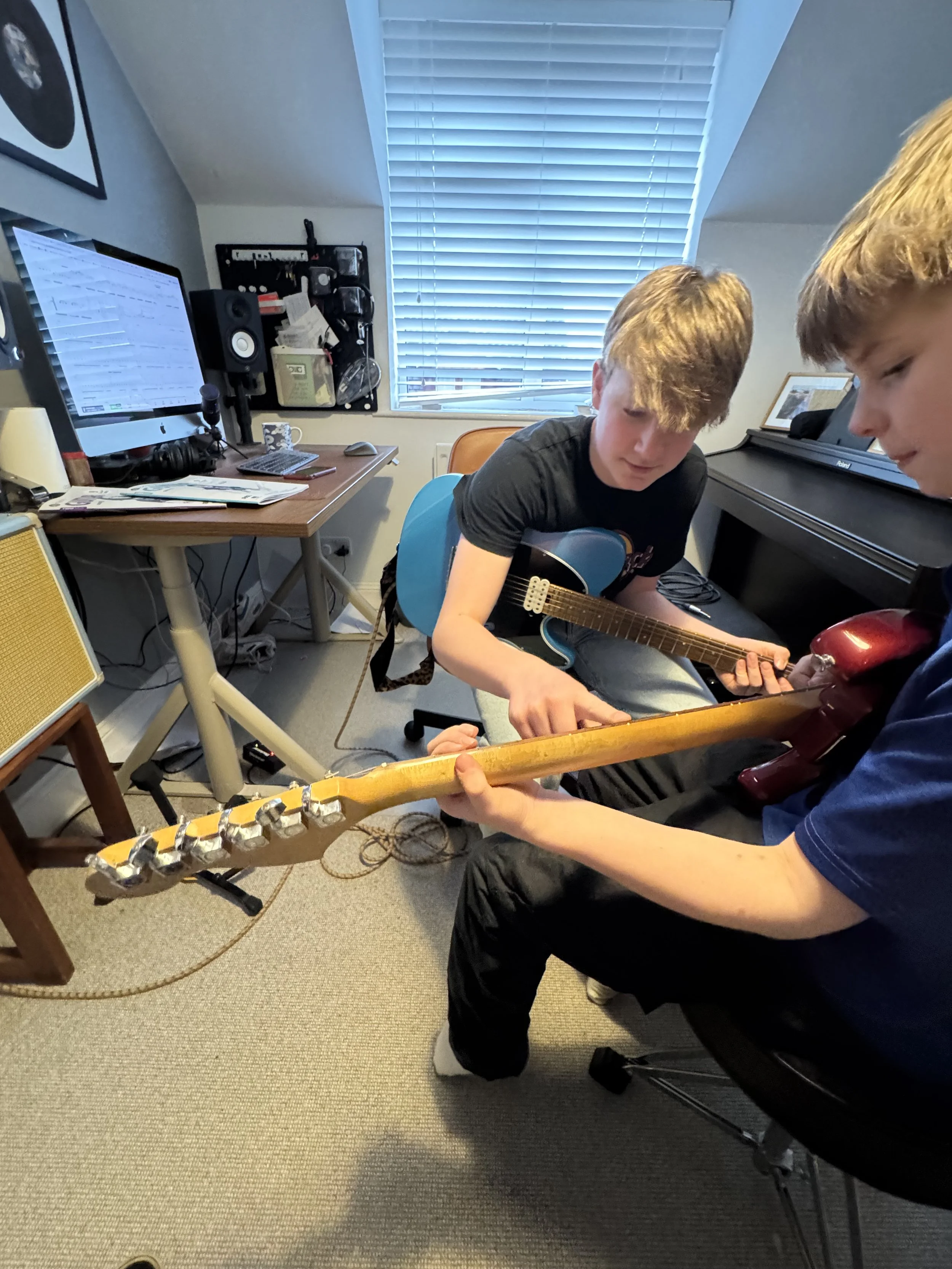Two young boys playing electric guitars in a music room. One boy with light-colored hair wearing a black shirt and the other with darker hair wearing a blue shirt. A computer and music equipment are on the desk in the background.