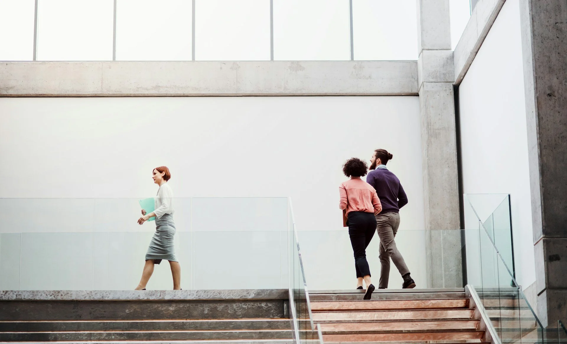 Three people walking up stairs in a modern building with large windows and concrete walls, one woman walking alone on the left, and a couple walking together on the right.