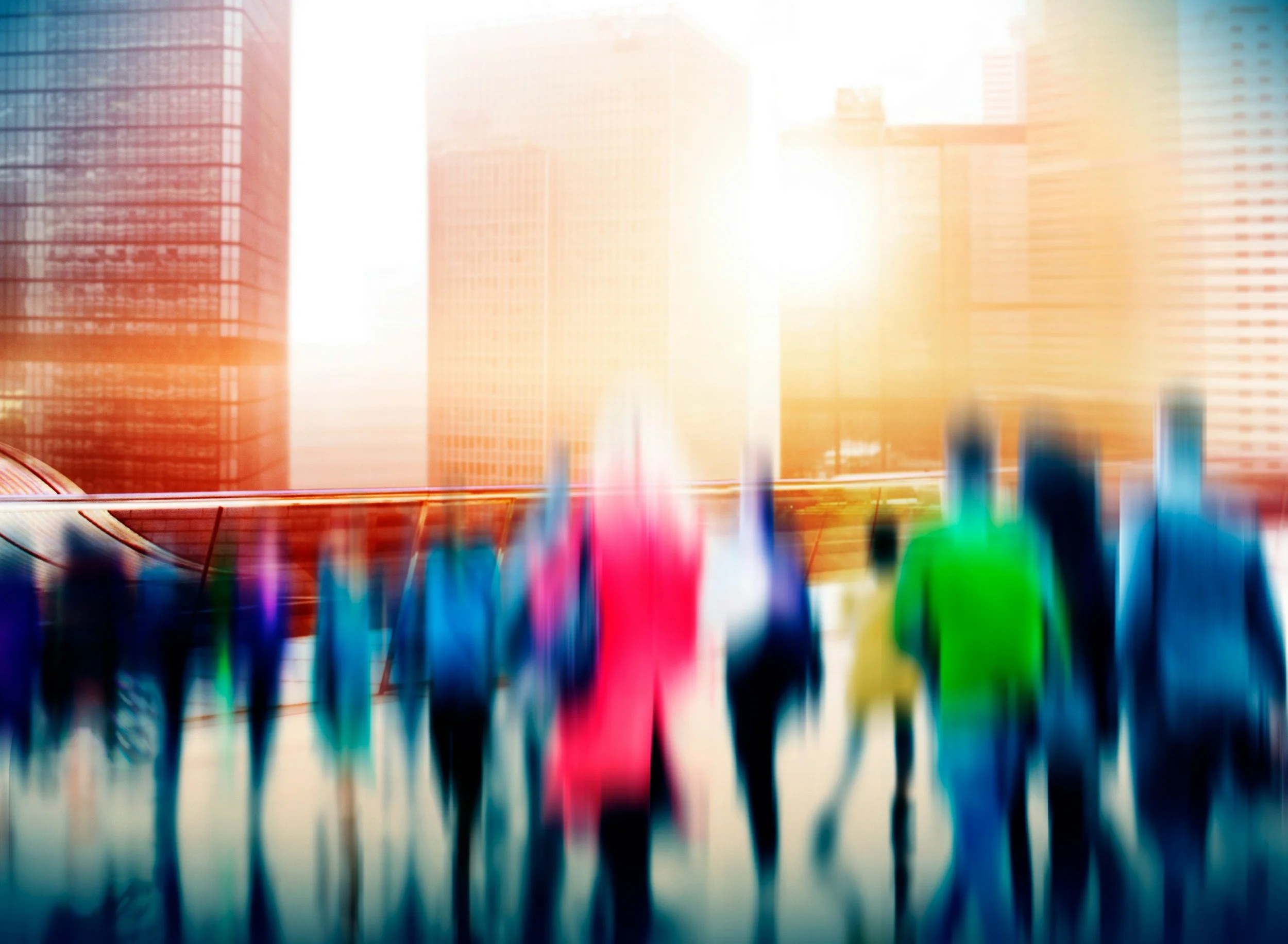 Blurred image of a crowd of people walking outdoors with tall buildings in the background and bright sunlight.