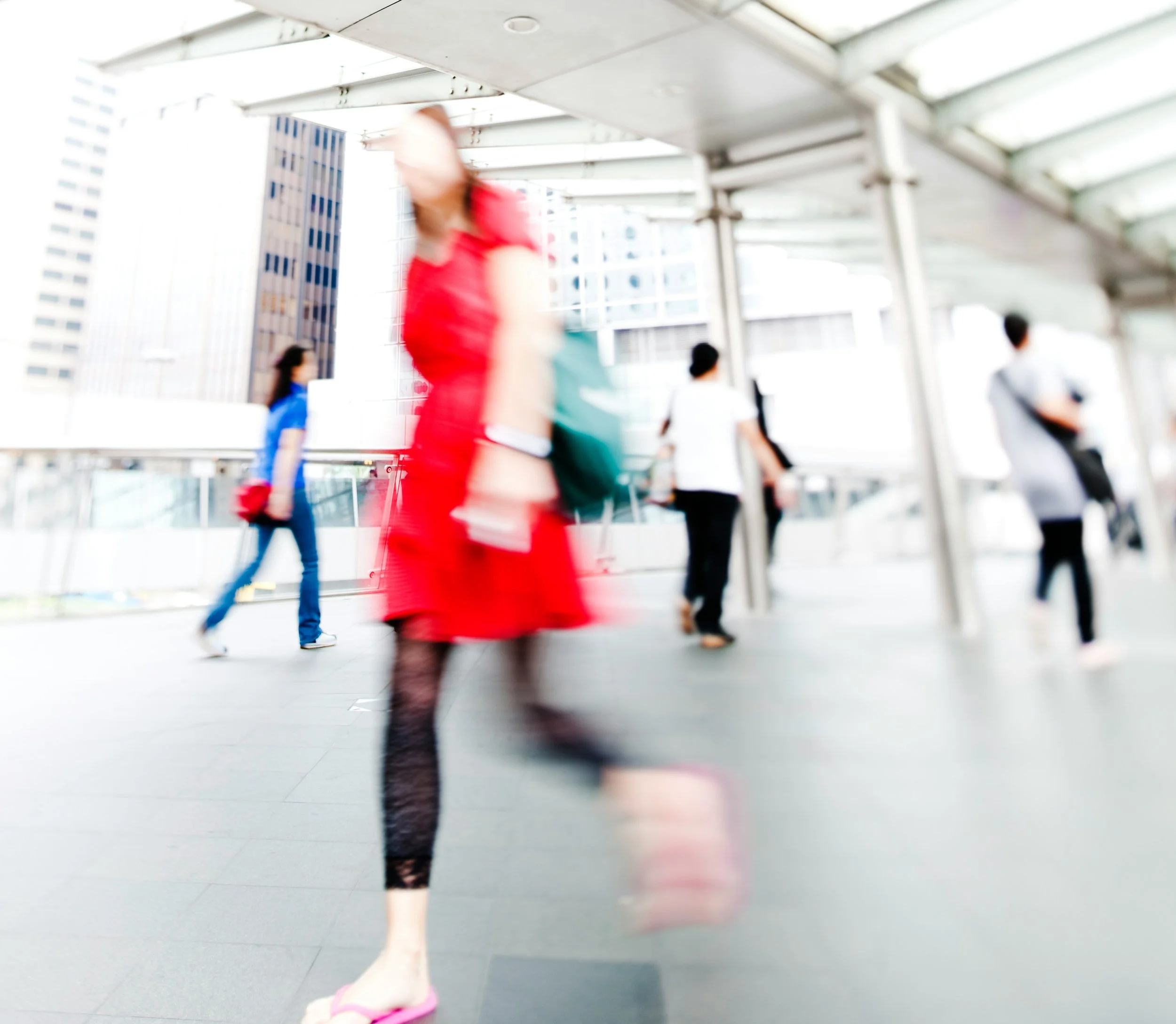 People walking on an outdoor covered walkway in an urban area, with tall buildings in the background. The photo is out of focus.
