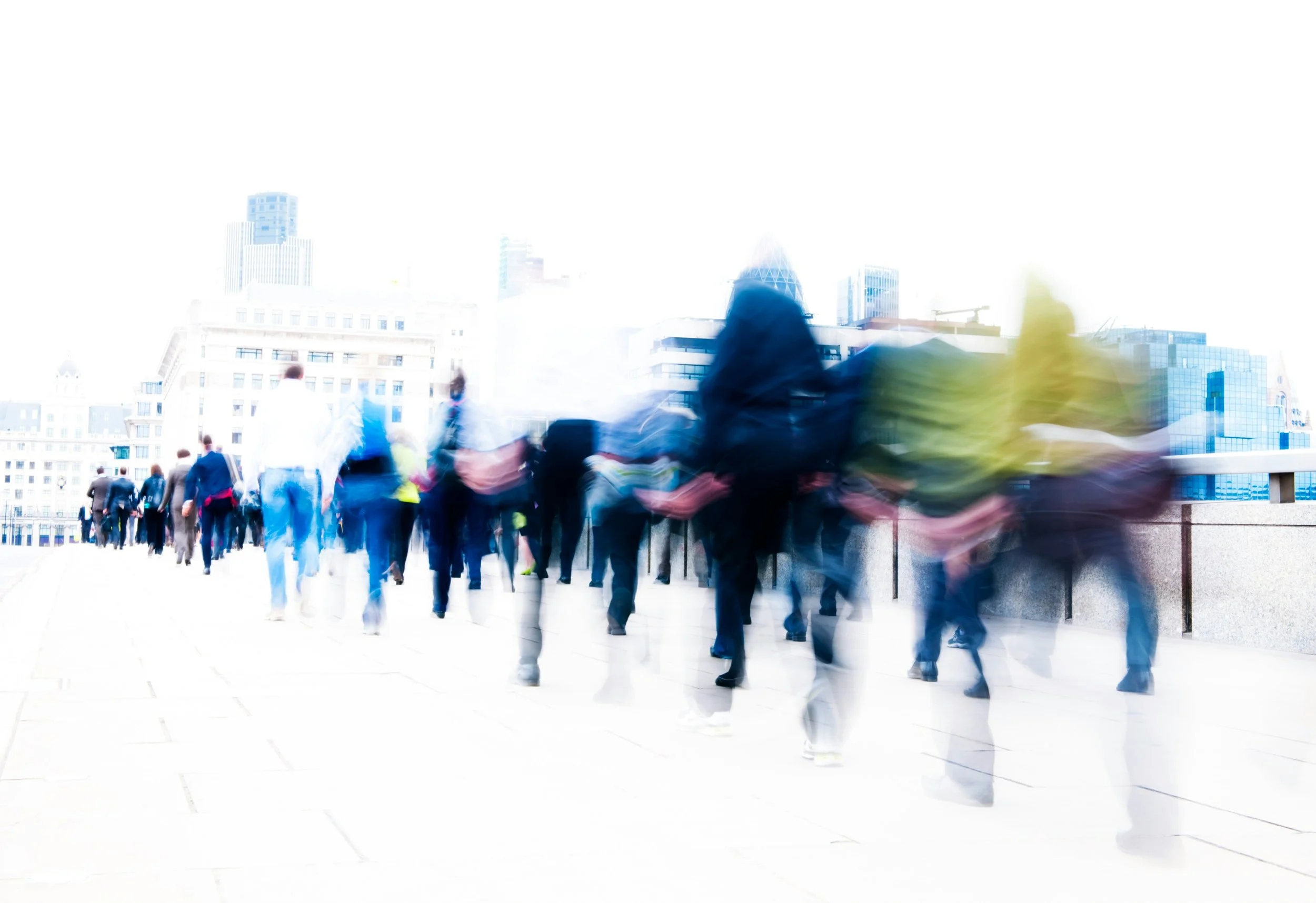Blurred crowd of people walking in a city during daytime, with tall buildings in the background.