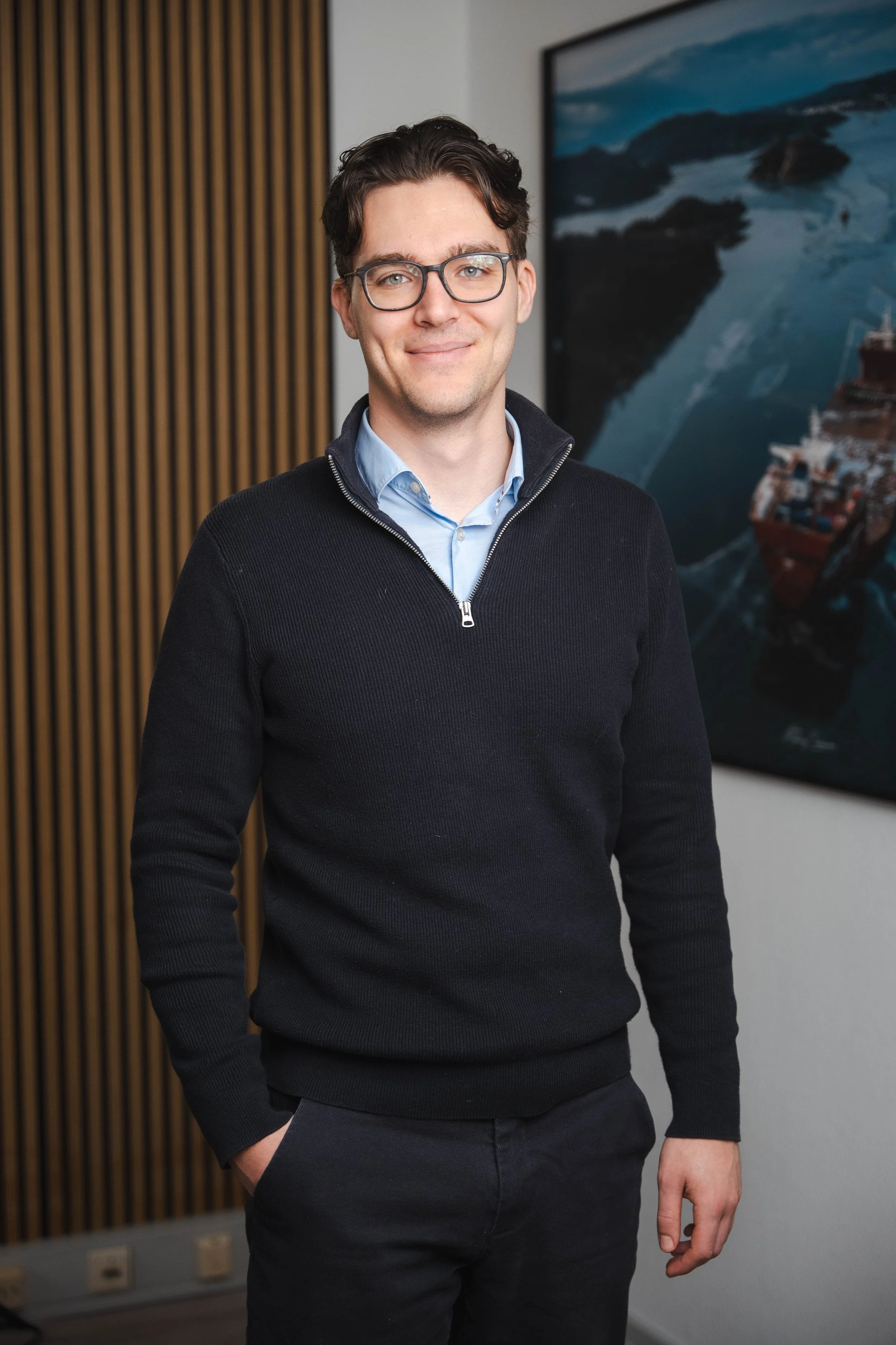 A young man with dark hair and glasses standing indoors, smiling, with a large landscape photograph of a river and a ship behind him.
