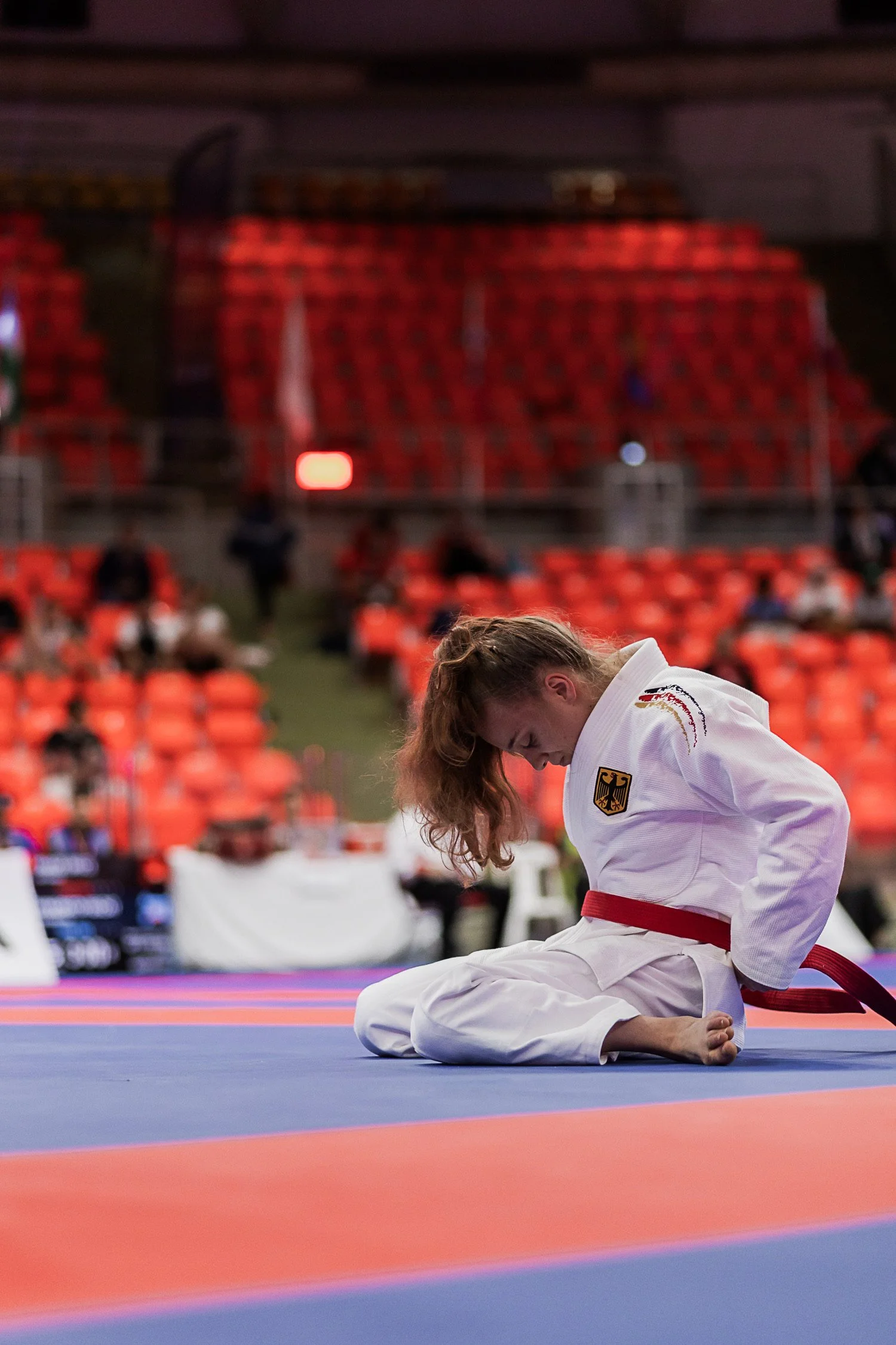 Junge Jiu-Jitsu-Kämpferin in weißem Gi mit rotem Gürtel in einer Turnhalle nach einem Match, sitzt auf dem Boden und sieht nach unten, im Hintergrund leere rote Sitzplätze.