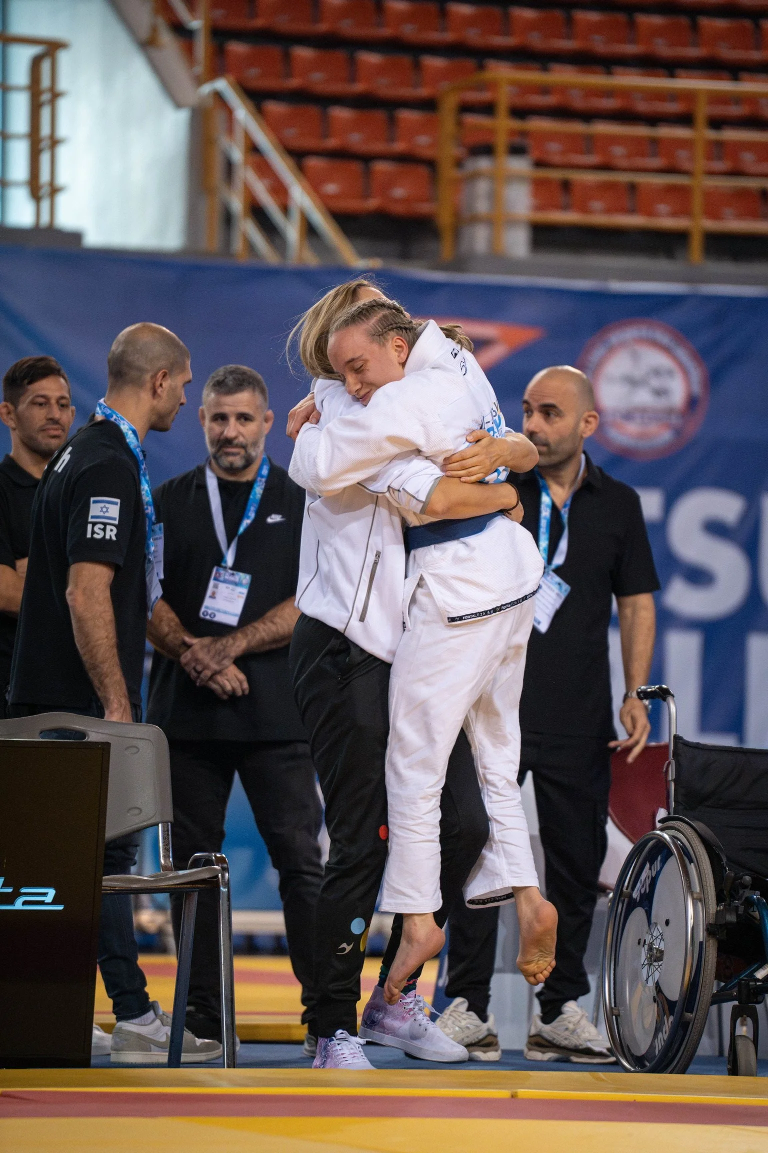 Zwei Frauen in Judoanzügen umarmen sich nach einem Wettkampf, im Hintergrund stehen Menschen mit Medaillen und einem Rollstuhl.