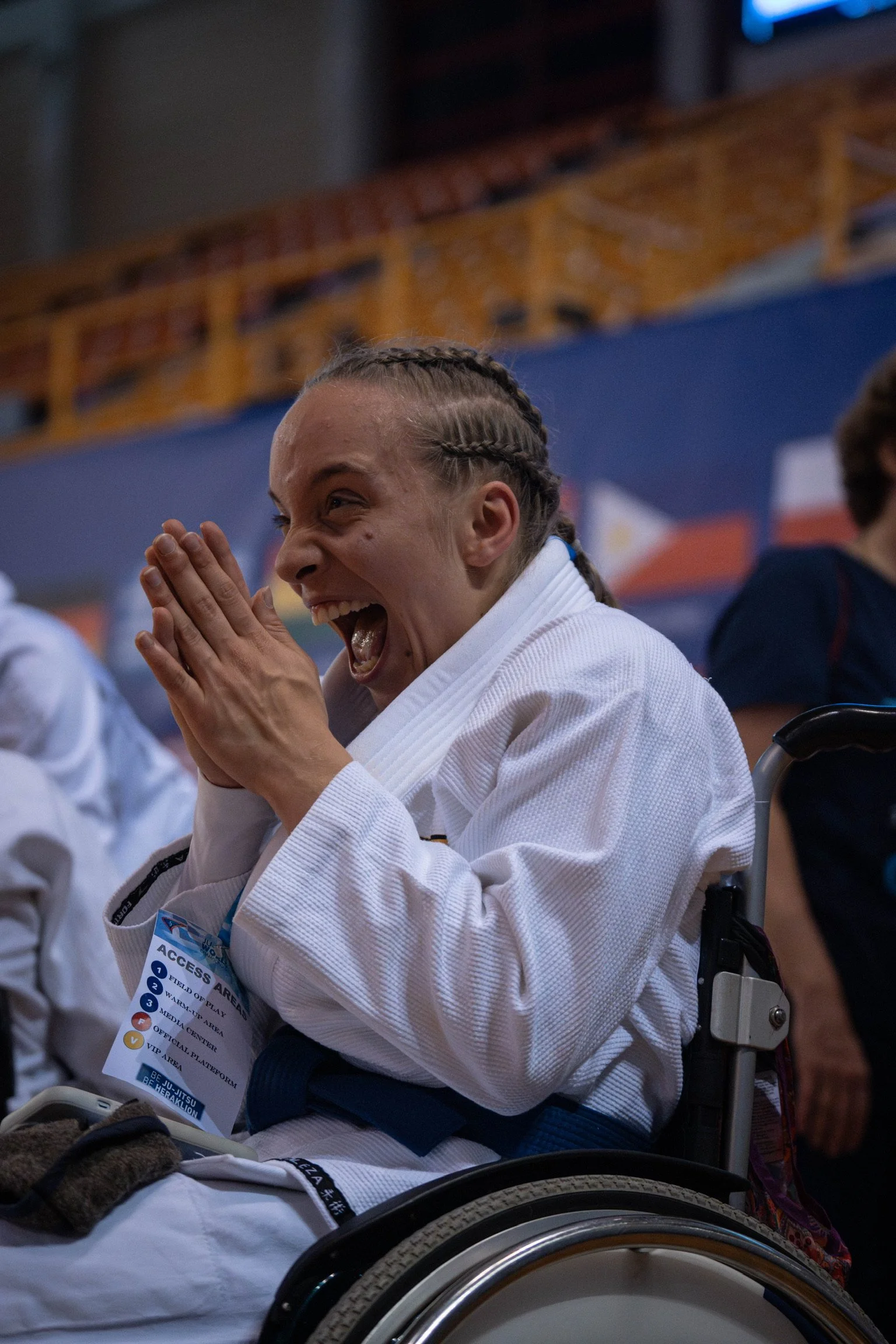 Ein lachender junge weibliche Taekwondo-Kämpferin in einem Rollstuhl, die die Hände vor dem Gesicht hält, bei einem Sportwettbewerb in einer Turnhalle.