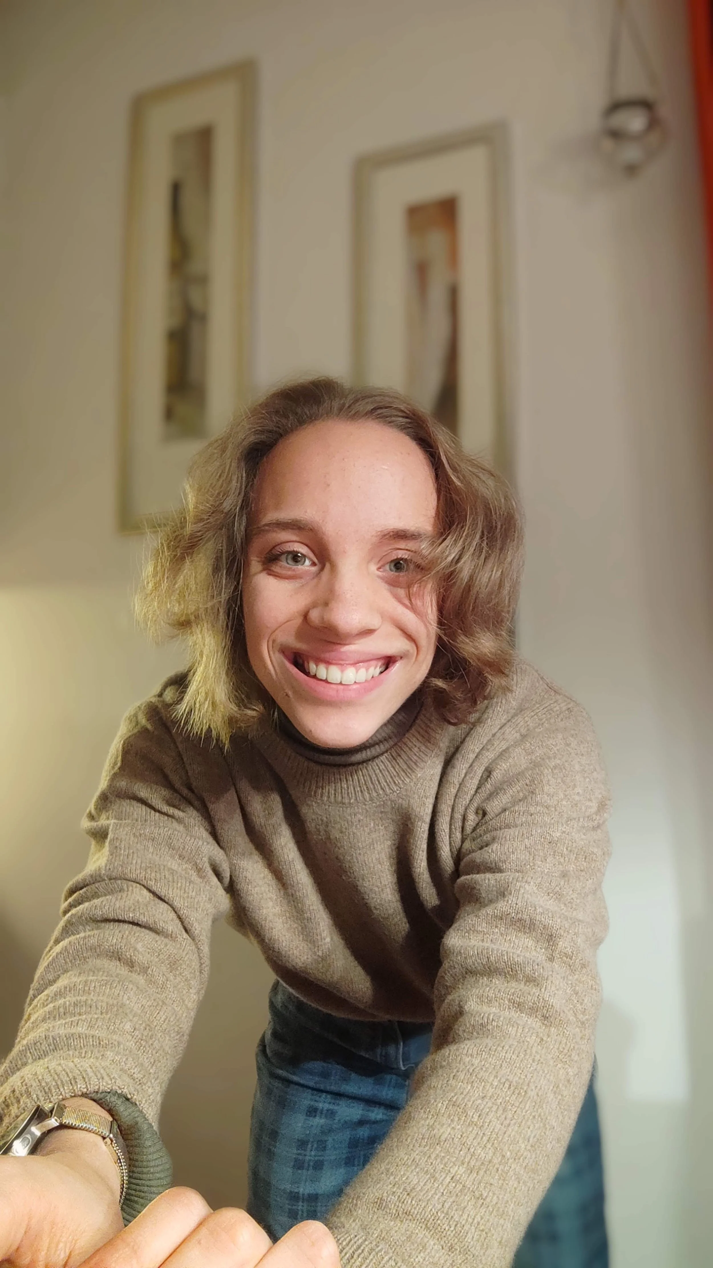 Junge Frau macht ein Selfie und lächelt, im Hintergrund sind Bilder an der Wand.