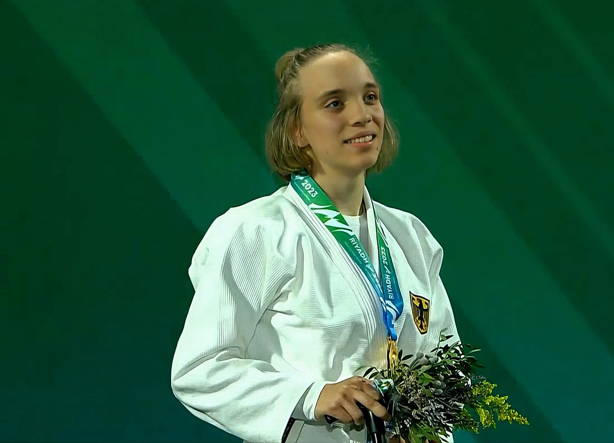 Eine junge weibliche Sportlerin im weißen Judo-Gi mit Deutschland-Emblem, sie trägt eine Goldmedaille um den Hals und hält einen Blumenstrauß, während sie auf einem Podest steht.