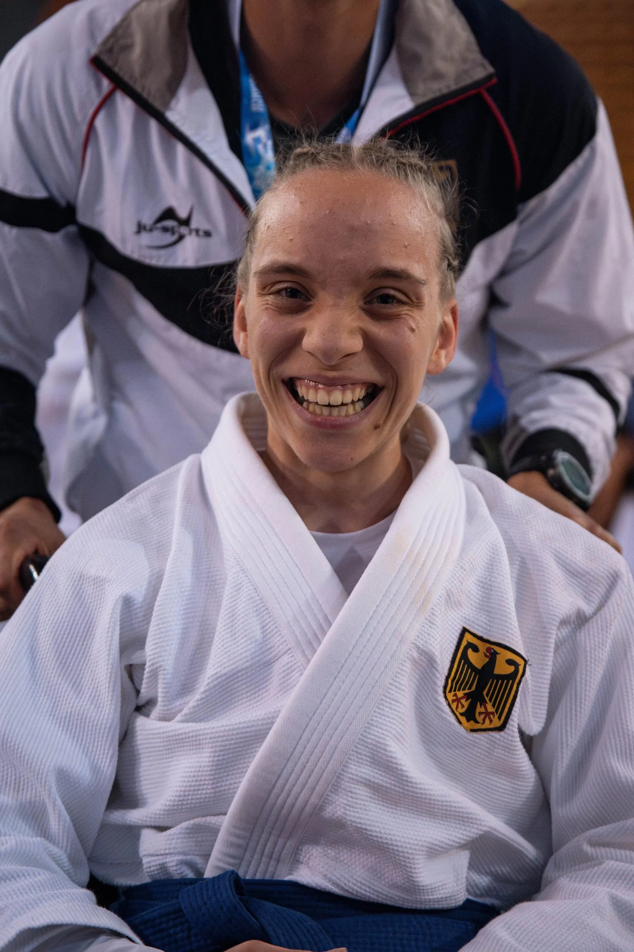 Eine lachende junge Frau in einem weißen Judo-Gi mit deutschem Wappen auf der Brust, im Hintergrund ein Mann in Wettkampfkleidung.