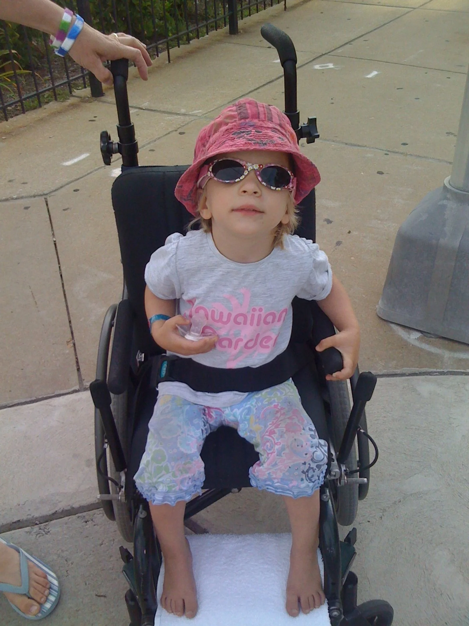 Ein Kind in einem Rollstuhl trägt eine rote Hut und Sonnenbrille, hält ein Spielzeug und sitzt auf einem weißen Handtuch auf dem Rollstuhl. Es ist warm und sonnig im Freien.