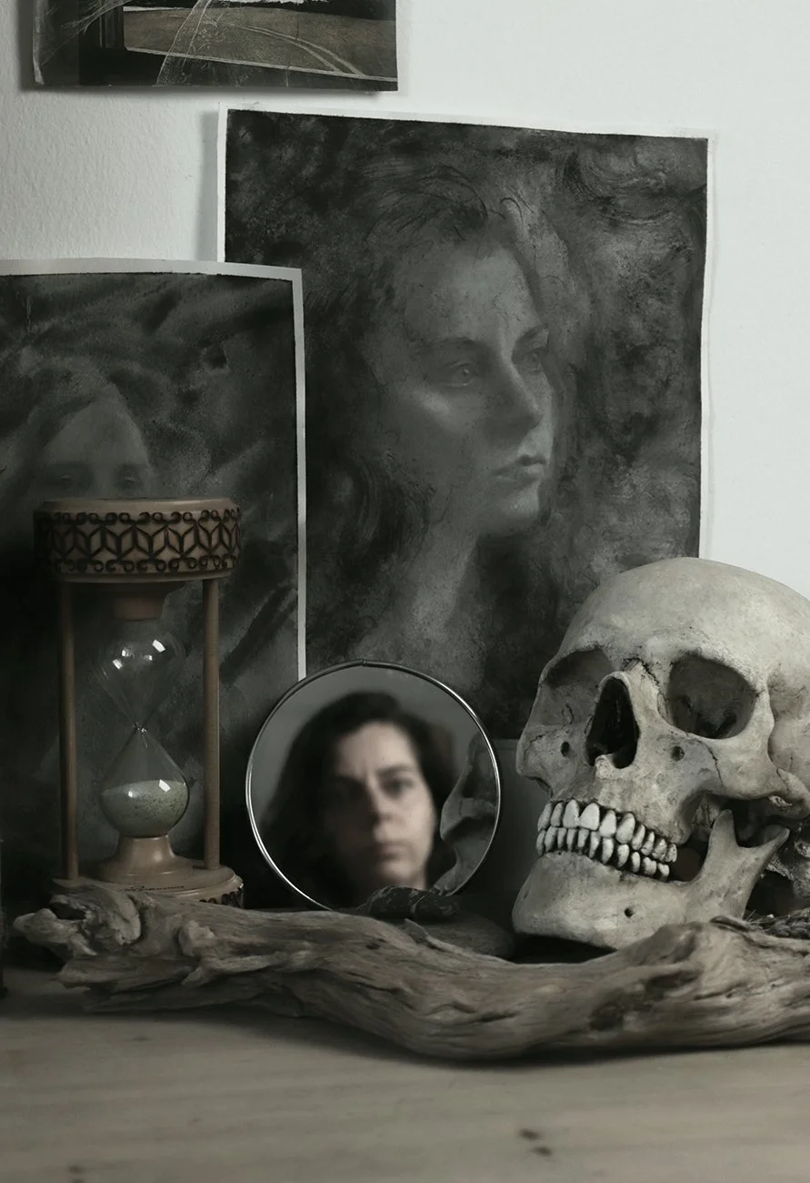 A collection of items including a human skull, a small round mirror reflecting a woman's face, an hourglass, and artworks of a woman's face, set against a white wall and a wooden surface.