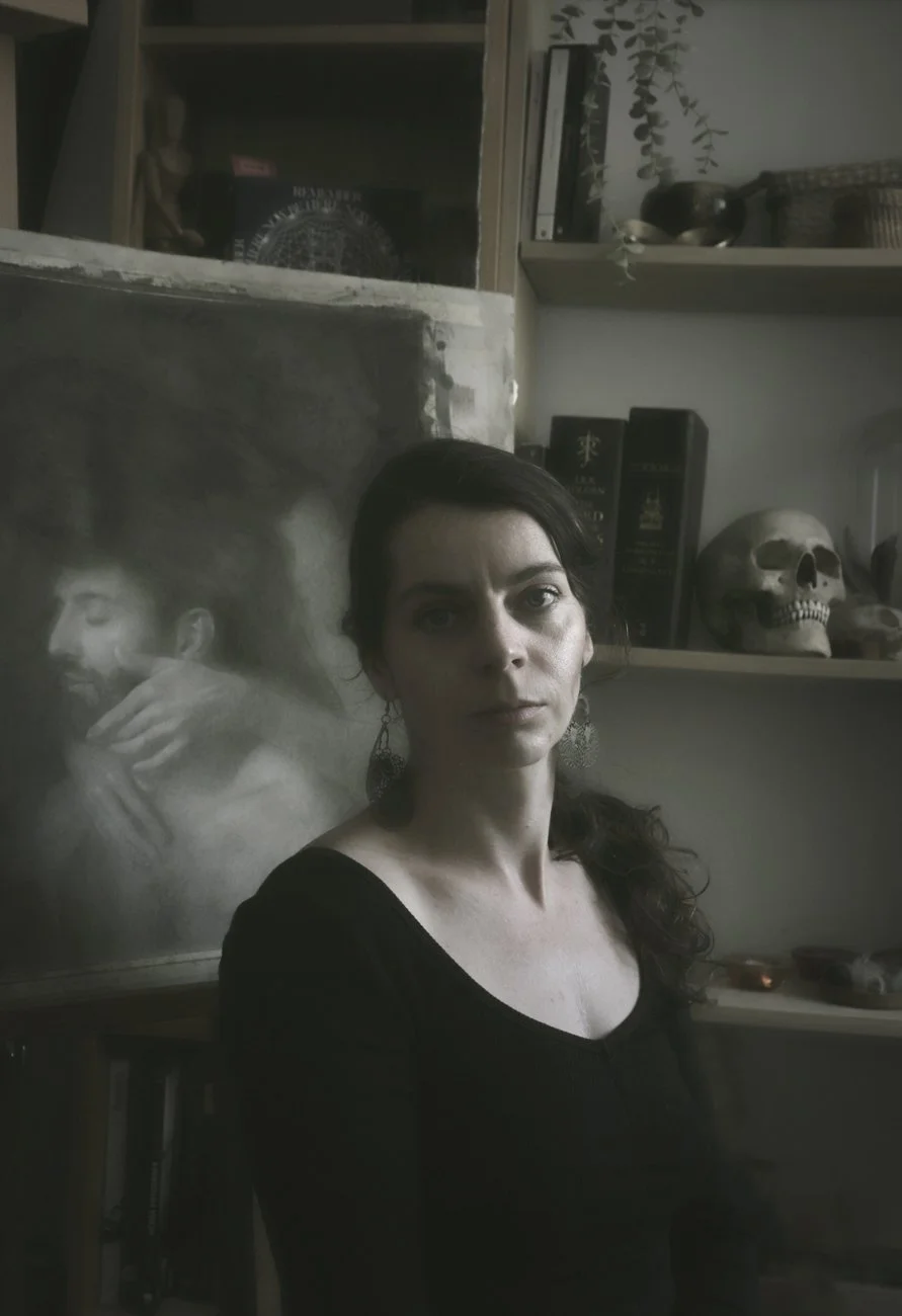 The artist Iva K with long dark hair and earrings standing in front of a shelf with skulls, books, and decorative items, with a painting of a man with a beard and mustache behind her.
