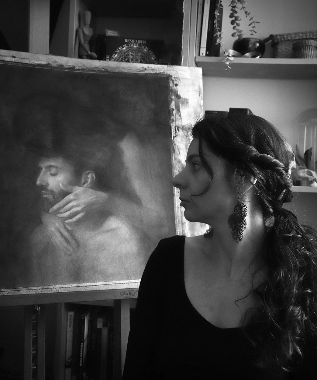 The artist Iva K with long, curly hair and large earrings looks at a black and white painting of a man with a beard, with a hand is resting on his neck.