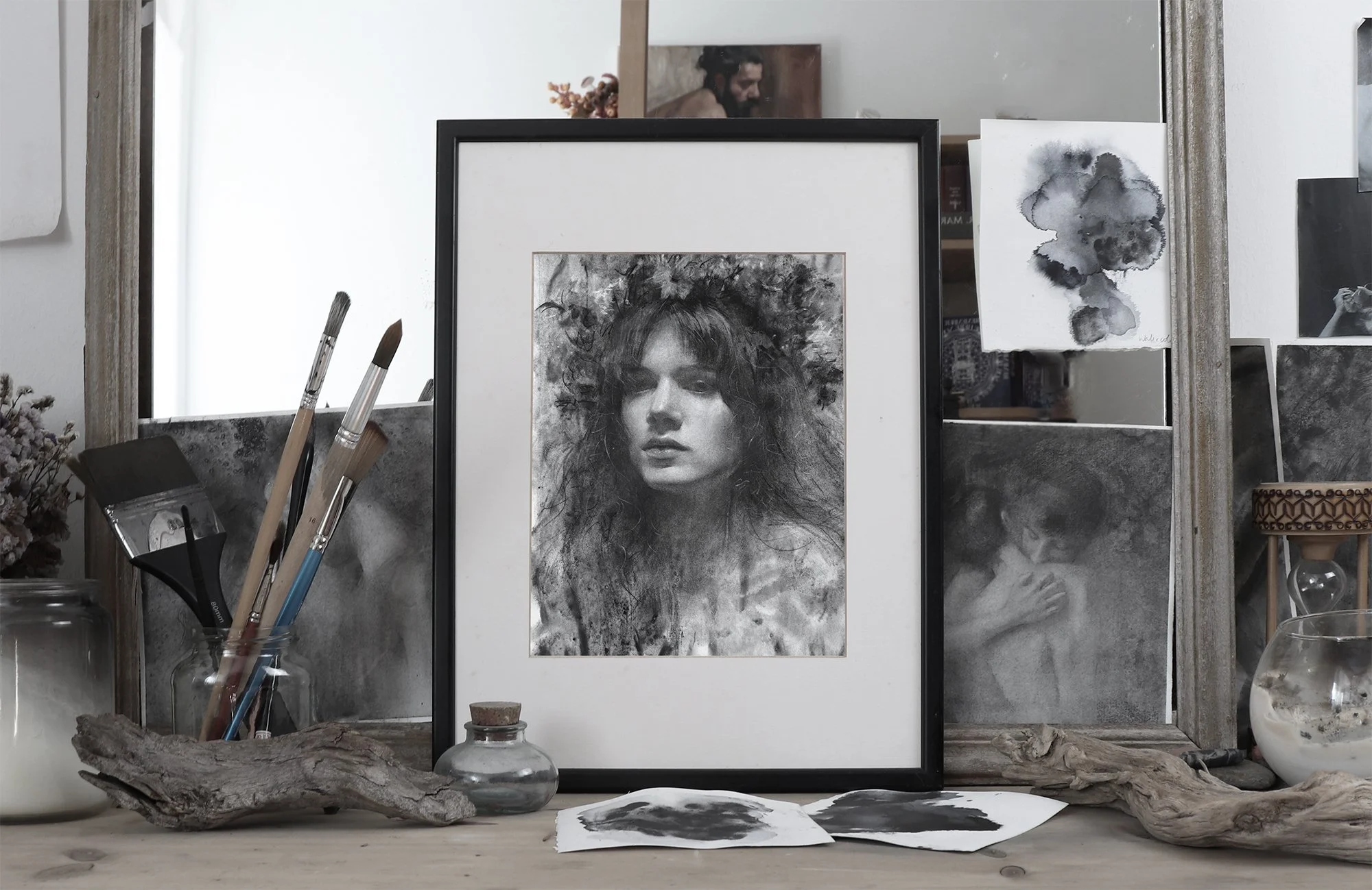 Art studio workspace with framed charcoal portrait of a woman with curly hair, a small sketch of a face, watercolor abstract art, paintbrushes in glass jar, and various art supplies on a wooden surface.