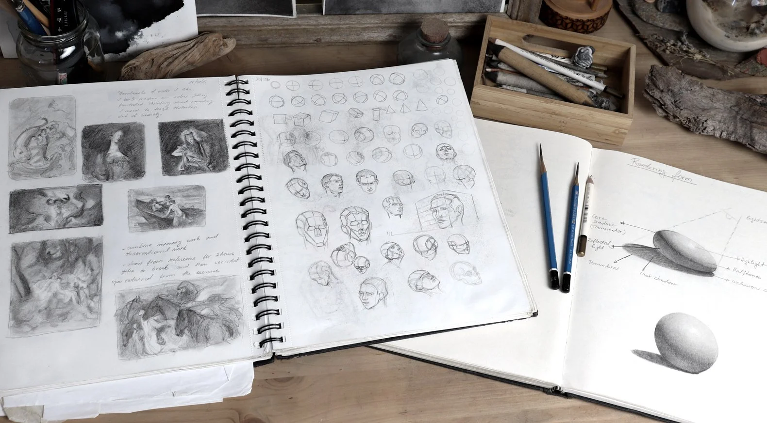 Open sketchbooks and drawing tools on a wooden table, with sketches of figures, skulls, and geometric shapes, including pencils and a wooden box with art supplies.