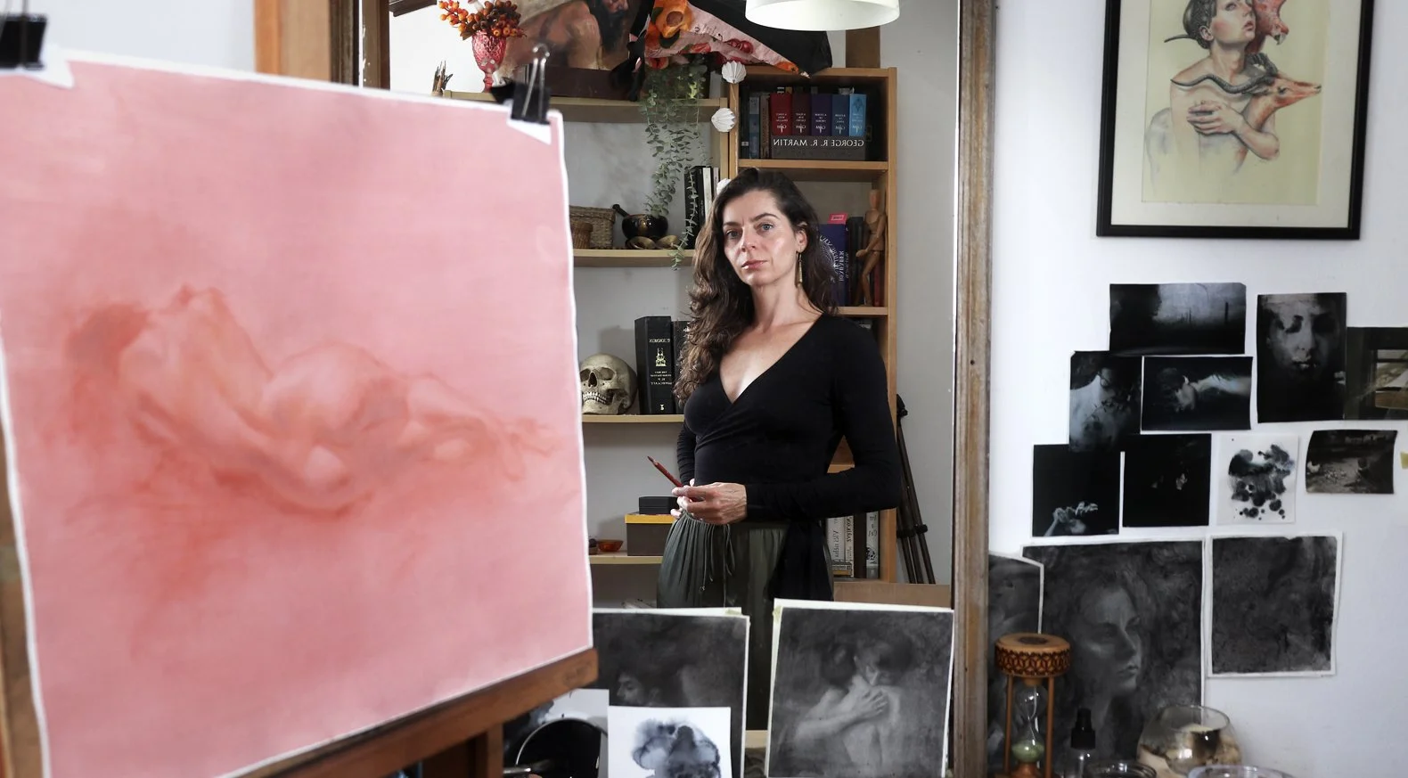 Artist Iva K  standing in an art studio surrounded by paintings and sketches, holding a paintbrush.