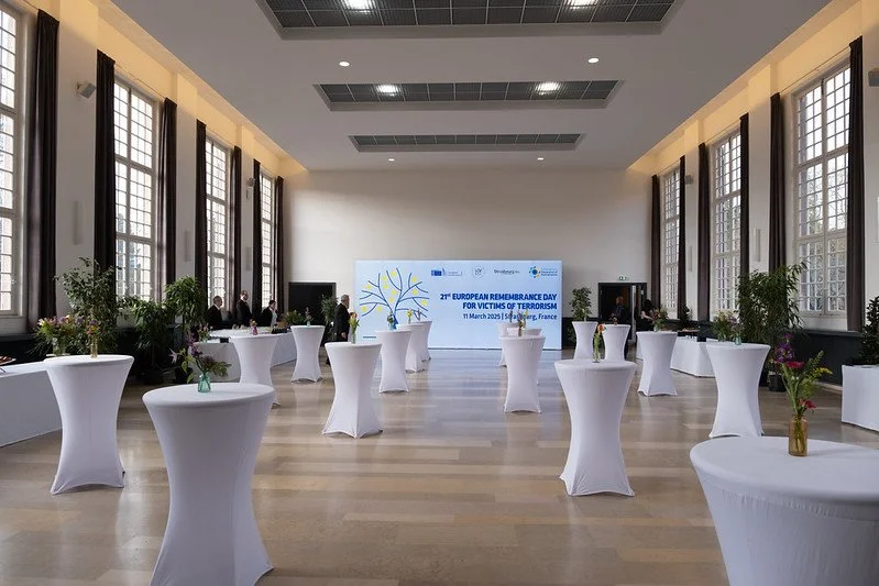 Large conference room with high ceilings and large windows on both sides. White round cocktail tables with vases of flowers arranged throughout. A large screen at the front displays the text "21st European Remembrance Day for Victims of Terrorism."
