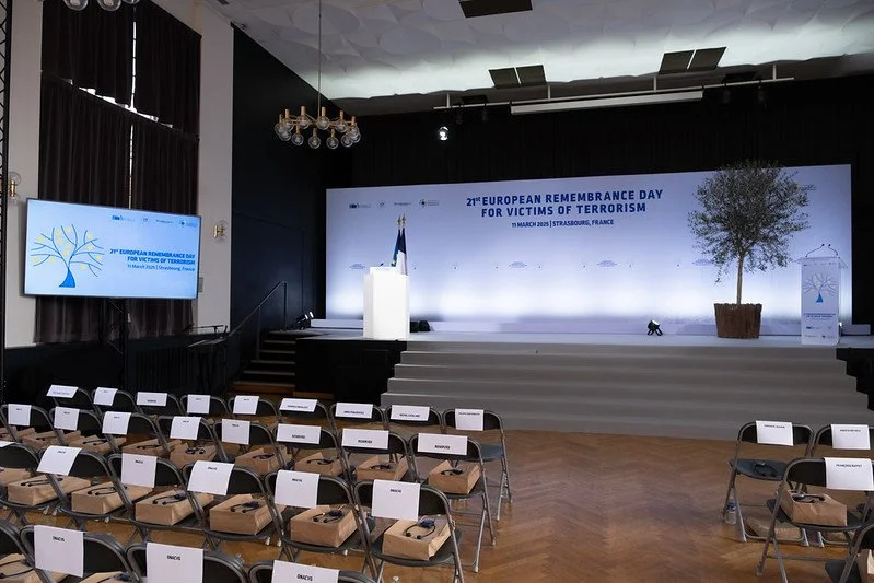 Conference room setup for European Remembrance Day event in France with chairs facing a stage, a large screen displaying event information, and a podium with flowers and flags.