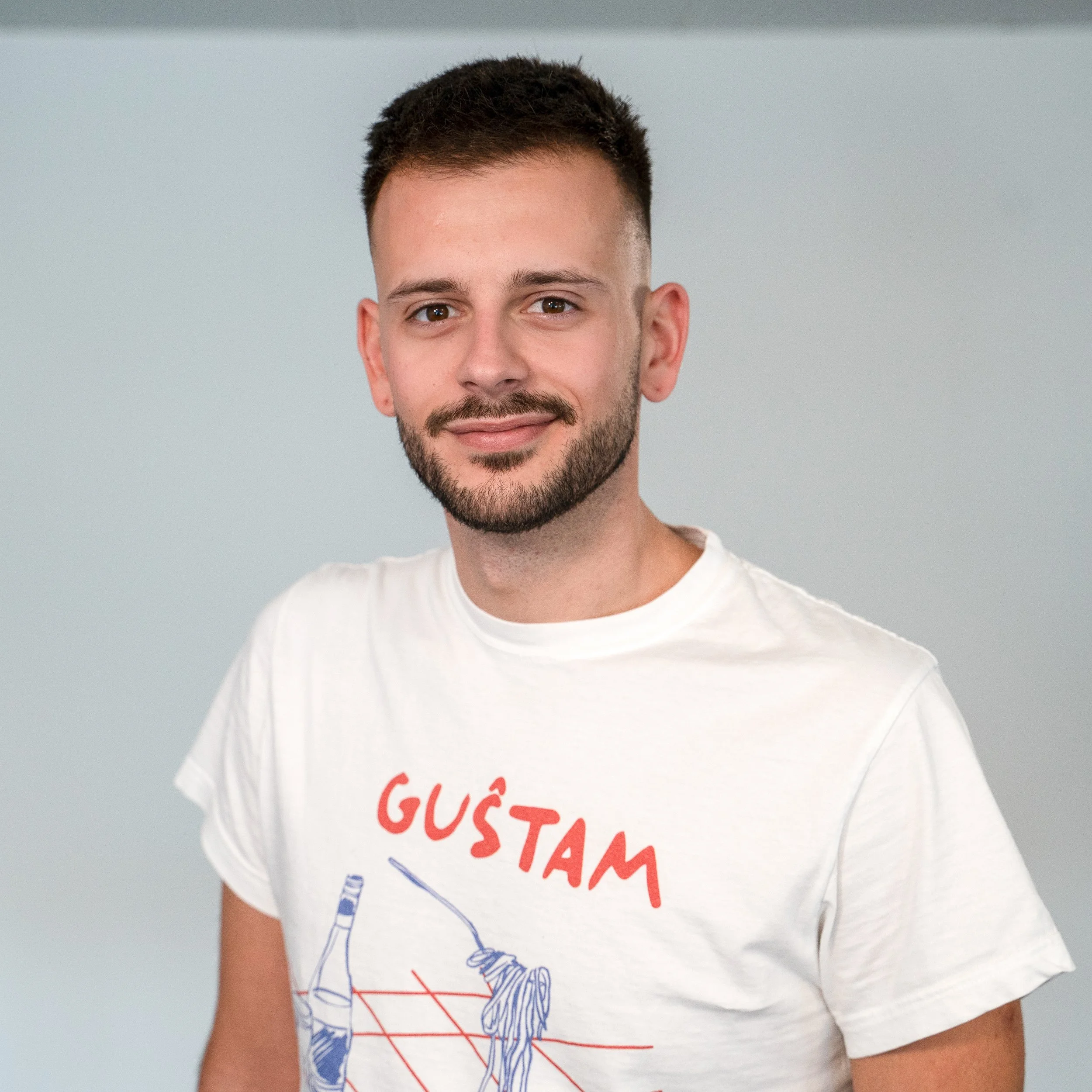 A young man with short dark hair and a beard, smiling, wearing a white t-shirt with red and blue graphics that include the word 'GUSTAM' and a bottle with a straw.