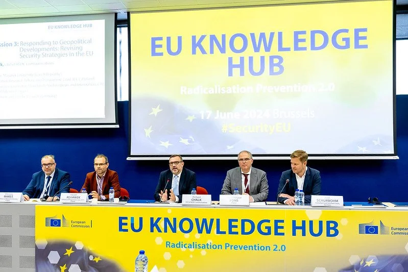 Panel of five men sitting at a long table during a conference, with a large screen behind them displaying "EU Knowledge Hub" and event details about Radicalisation Prevention 2.0 in Brussels on June 17, 2024.