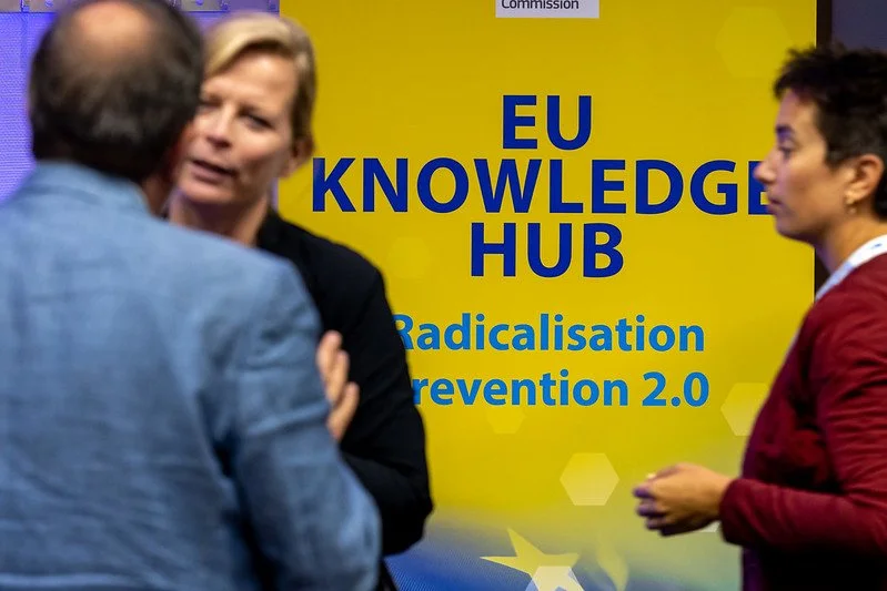 Three people are engaged in conversation in front of a yellow sign with blue text that reads 'EU Knowledge Hub: Radicalisation Prevention 2.0'.