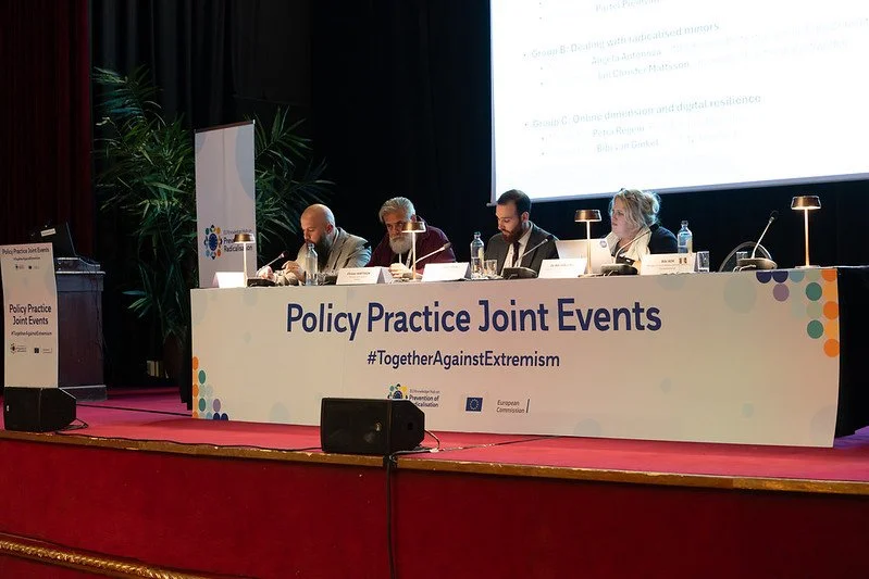 A panel of five people sitting at a long table on a stage, with a large screen behind them displaying presentation slides. The table has a banner with the text 'Policy Practice Joint Events #TogetherAgainstExtremism' and the European Commission logo.