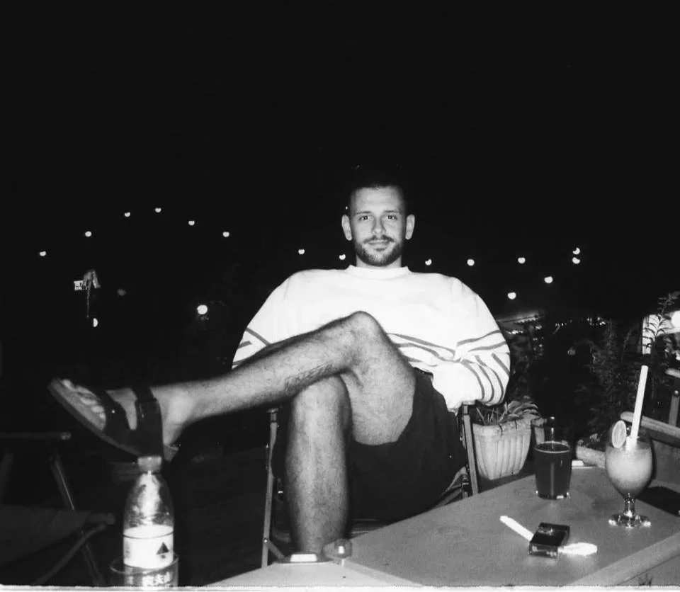 A man sitting at an outdoor table at night, with one leg crossed over the other, and drinks on the table.