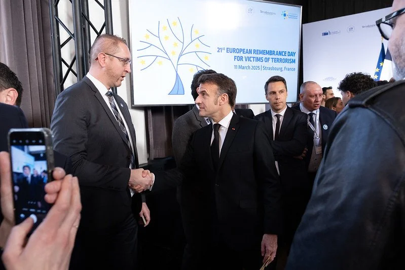 Two men in suits shaking hands at a conference, with a screen in the background displaying that it is the 21st European Remembrance Day for Victims of Terrorism on March 11, 2025, in Strasbourg, France.
