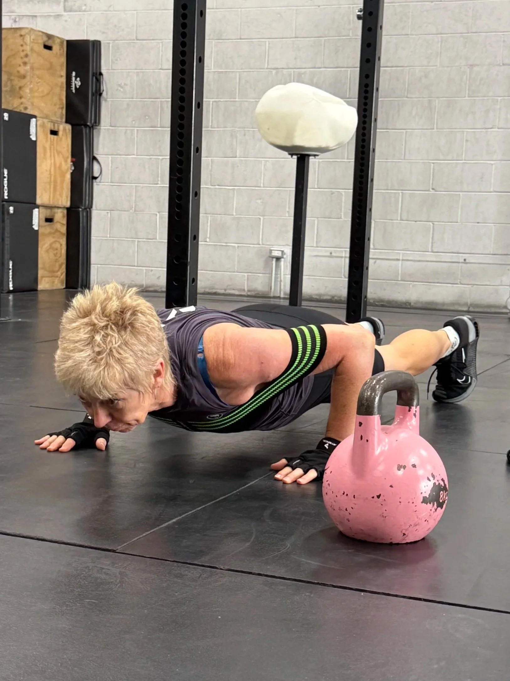 An older woman doing a push-up in a gym, with a kettlebell nearby. She is wearing a black workout outfit and fingerless gloves.
