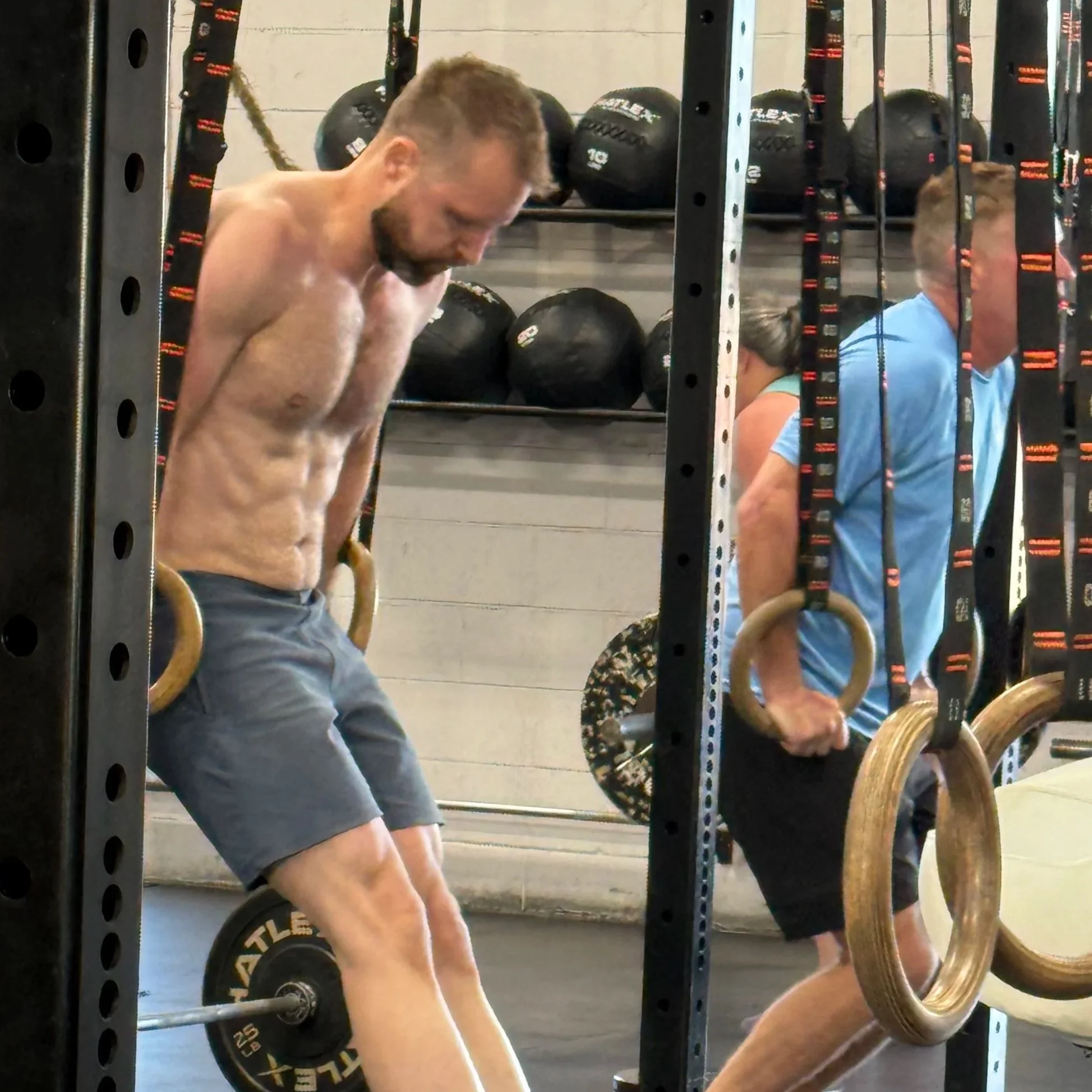 Two men with muscular builds and short hair are doing pull-ups on gym rings in a gym, with black kettlebells and shelves in the background.