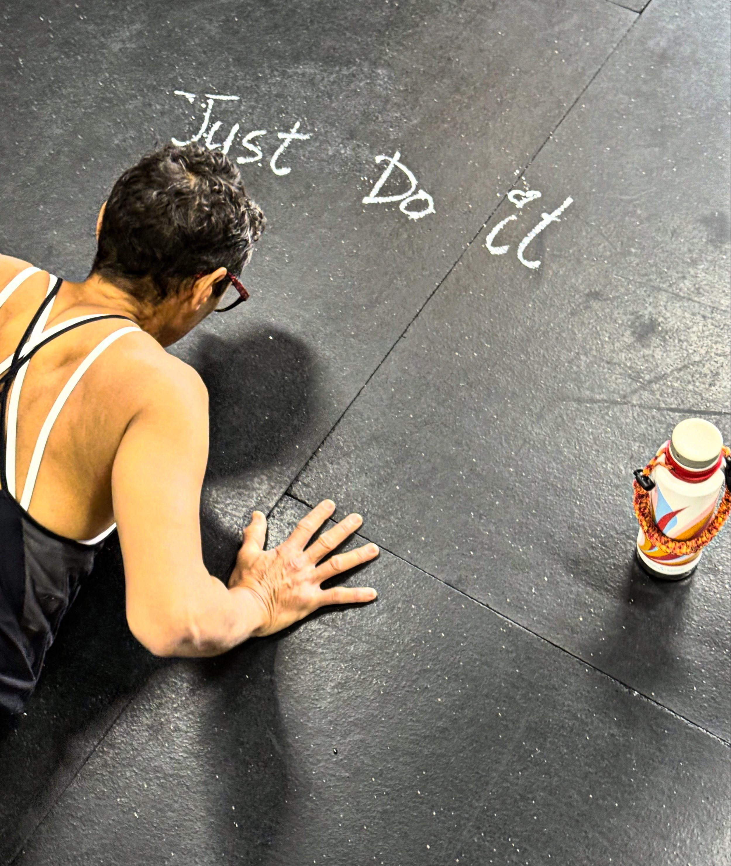 A woman in fitness attire doing a push-up on a gym floor, with the words 'Just Do It' written in chalk on the floor next to her and a water bottle nearby.