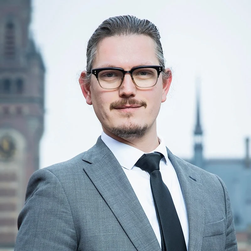 A man in a gray suit, white shirt, and black tie with glasses standing outdoors in front of a cityscape with historic buildings.
