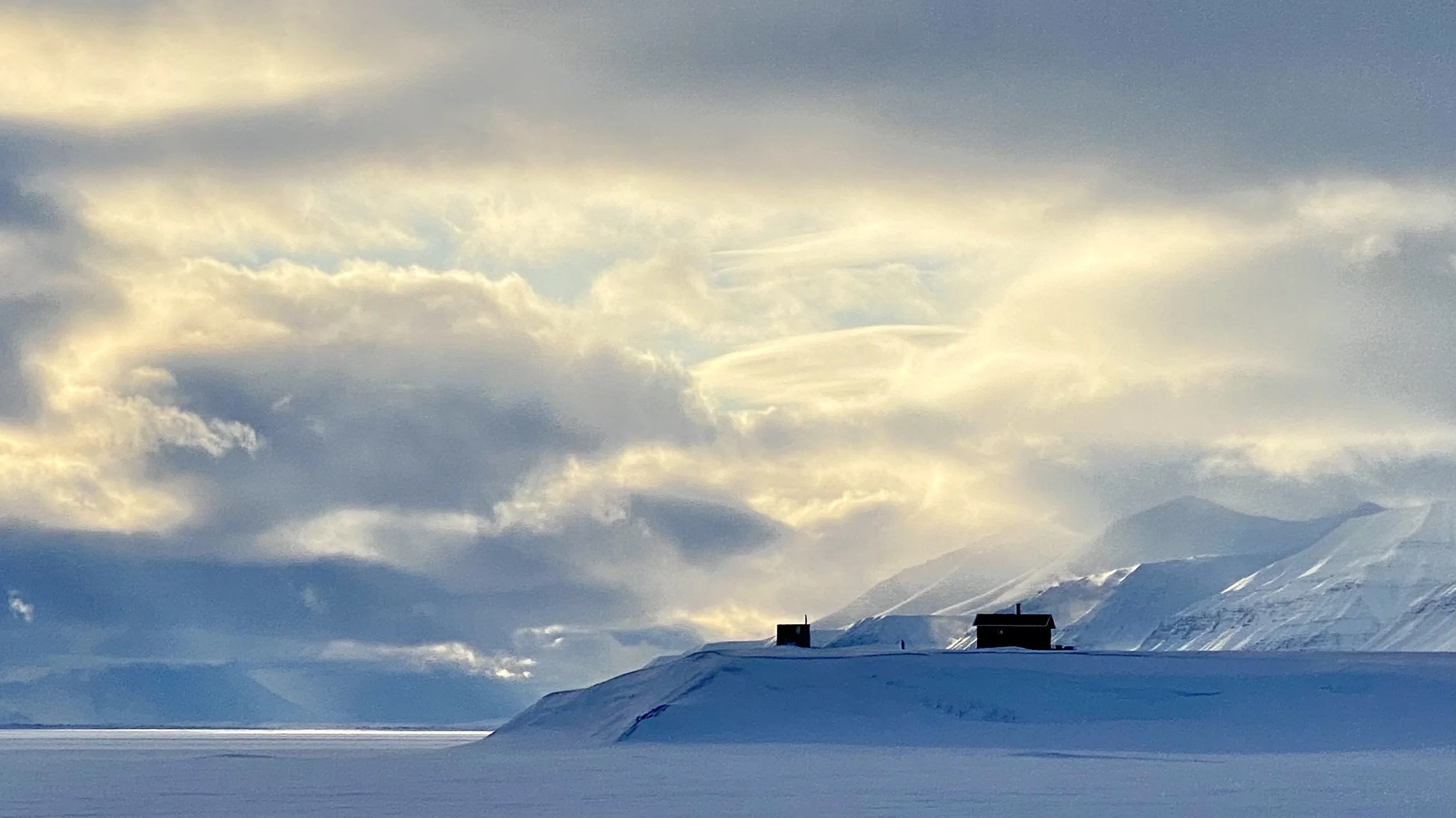 Snow-covered landscape in remote, high arctic, with two small buildings on a hill, mountains in the background, under a cloudy sky. Quiet, reflective, solitary, unique, offering space to think away from the world.