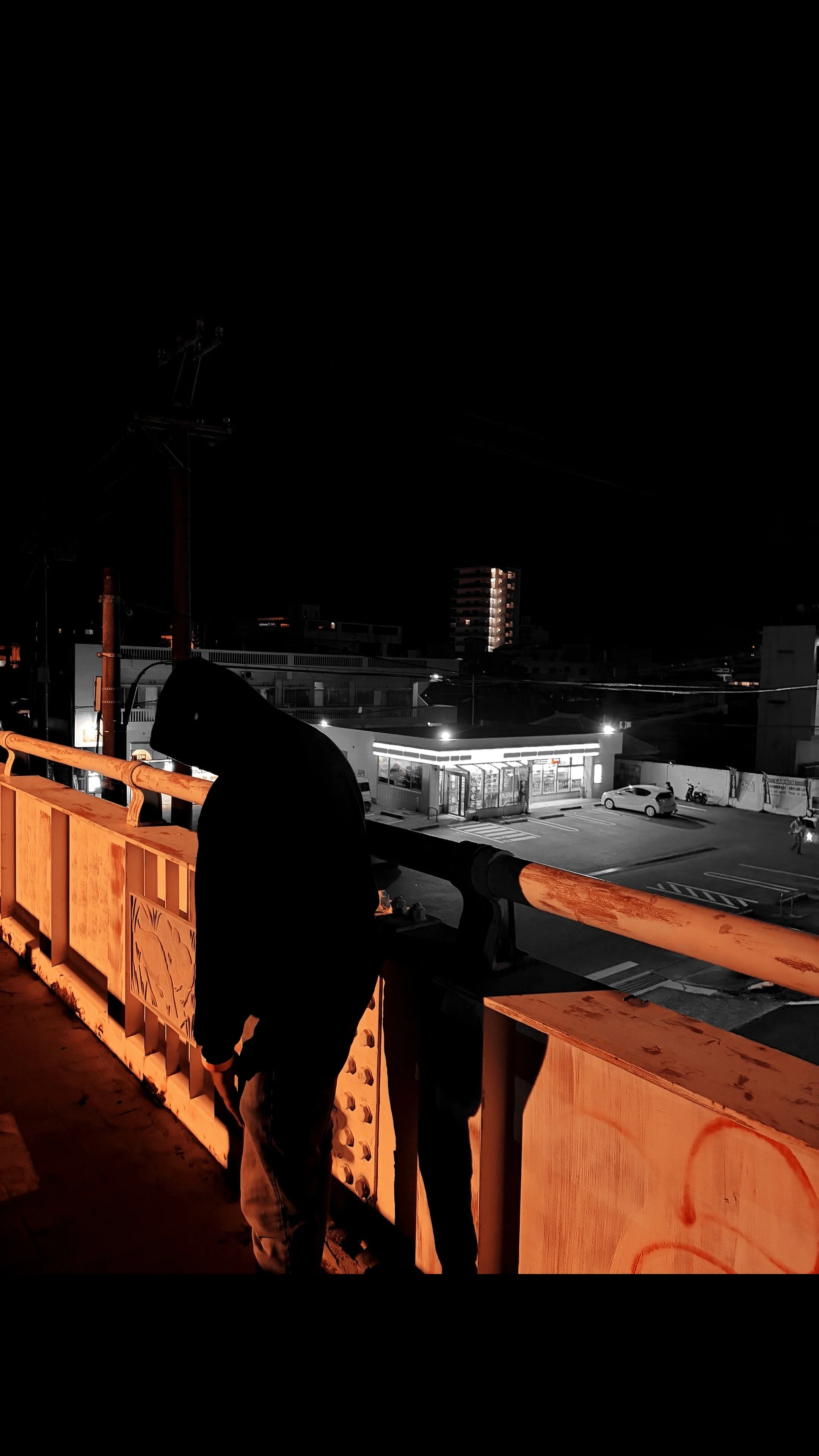 Moody nighttime city scene with a hooded NOIR$ standing on an overpass, overlooking an urban street illuminated by streetlights