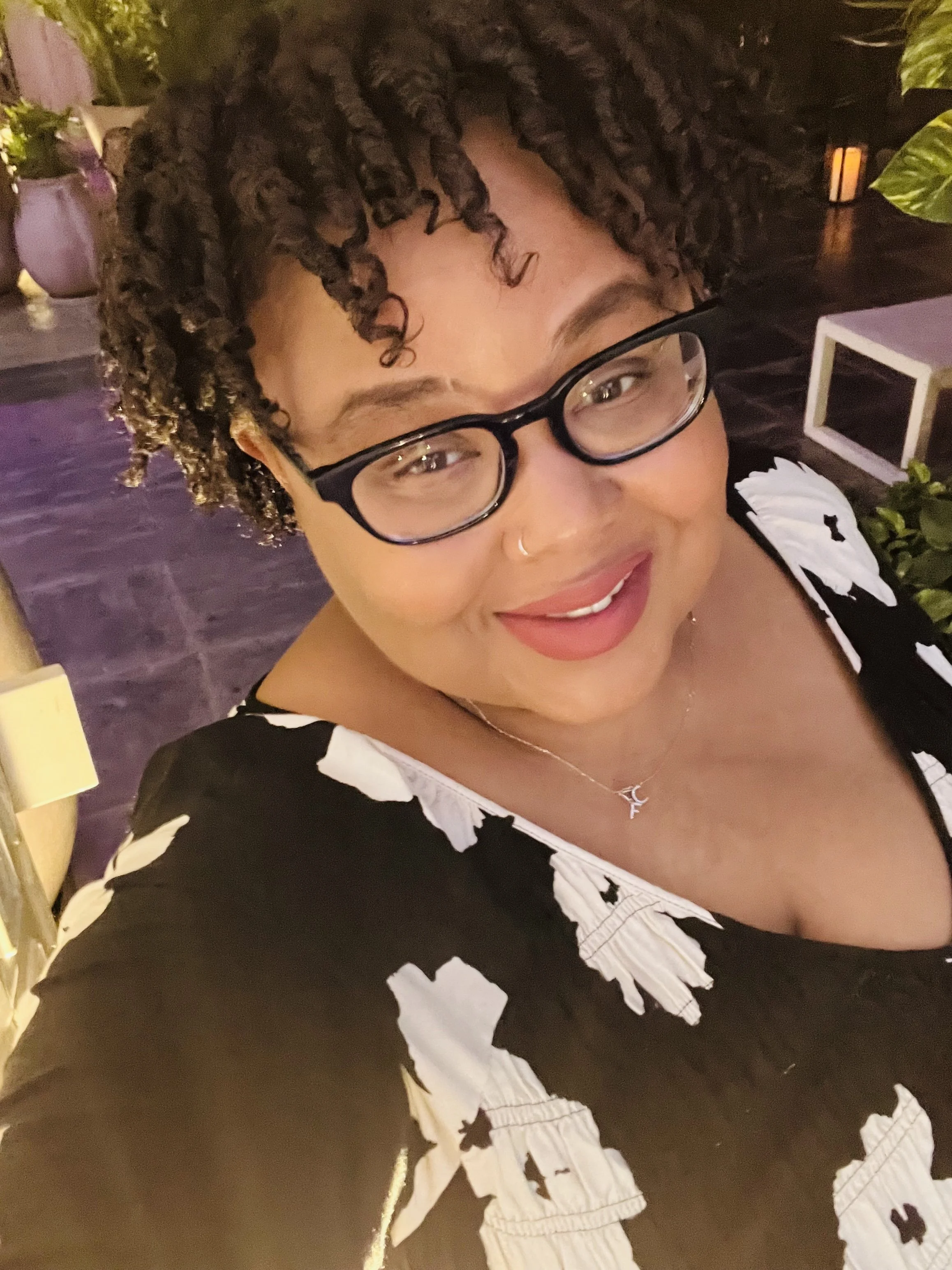 A smiling woman with curly hair, glasses, a nose ring, and a necklace taking a selfie indoors. She is wearing a black top with a white pattern and is surrounded by plants and soft lighting.