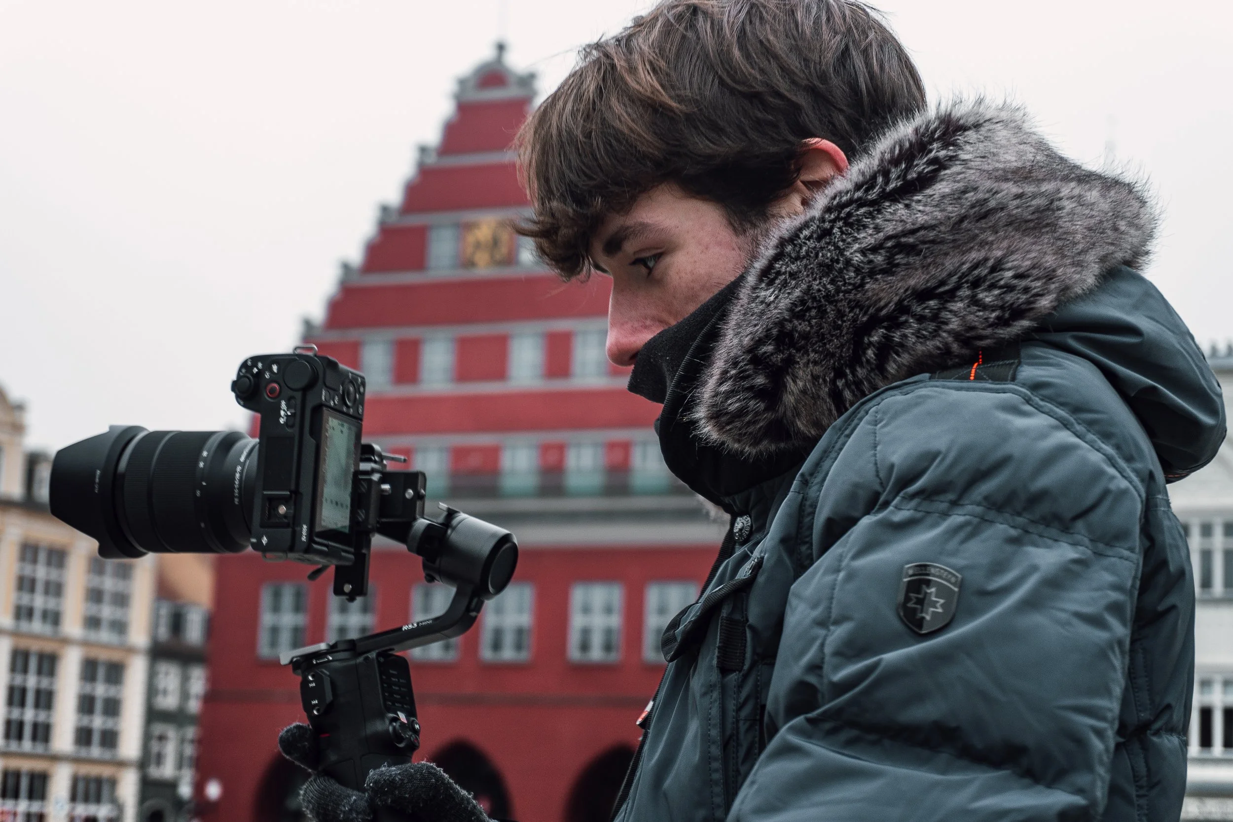 Junge Person mit dunklem, mehrfarbigem Wintermantel mit Fellkapuze, hält eine Kamera mit Gimbal in der Hand vor einer roten Gebäudefront in einem urbanen Umfeld.