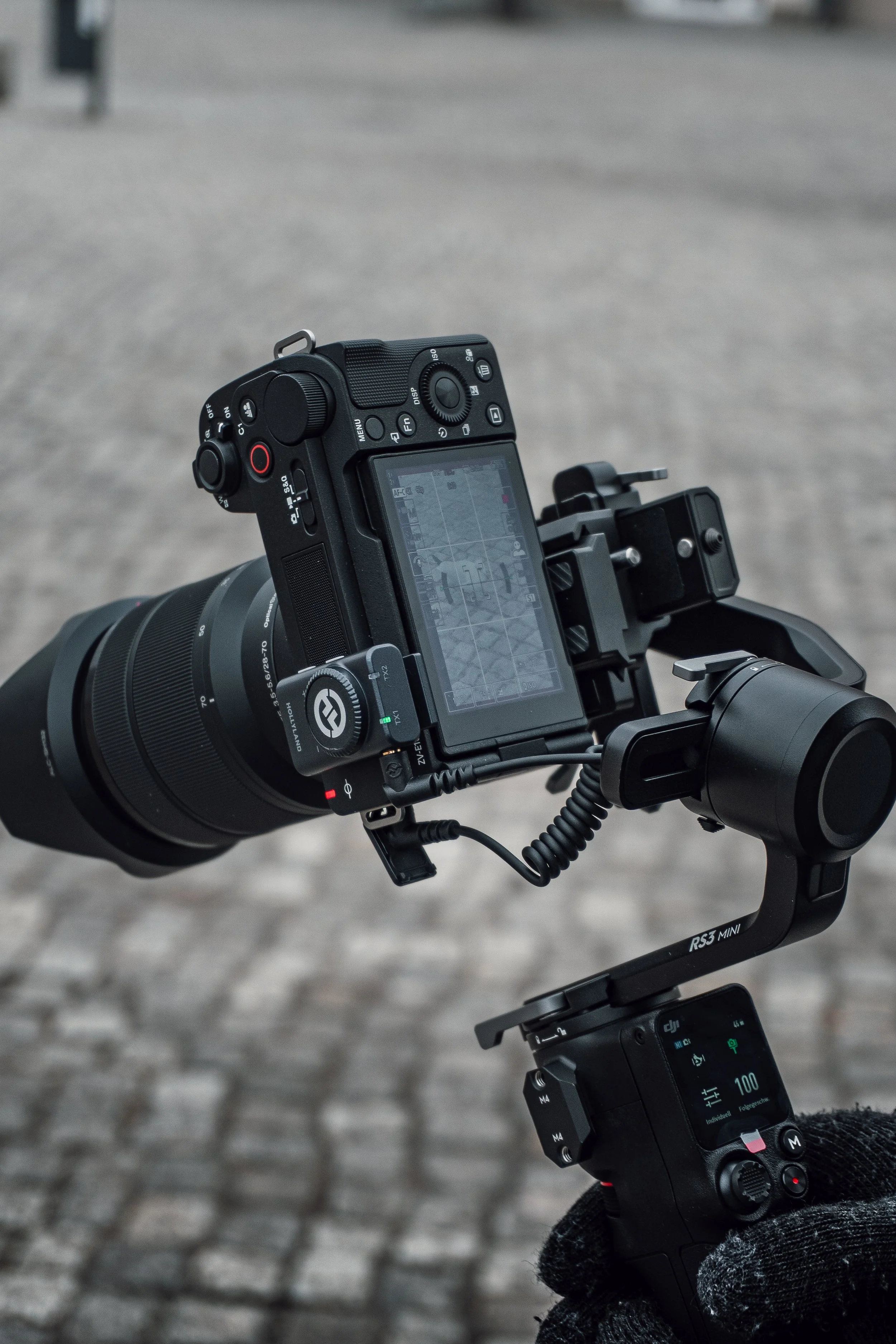 Aufnahmegerät mit Kamera auf einer ländlichen Straße, mit Steuerungsknöpfen und einem Bildschirm, beobachtet von einer Person mit schwarzem Handschuh.