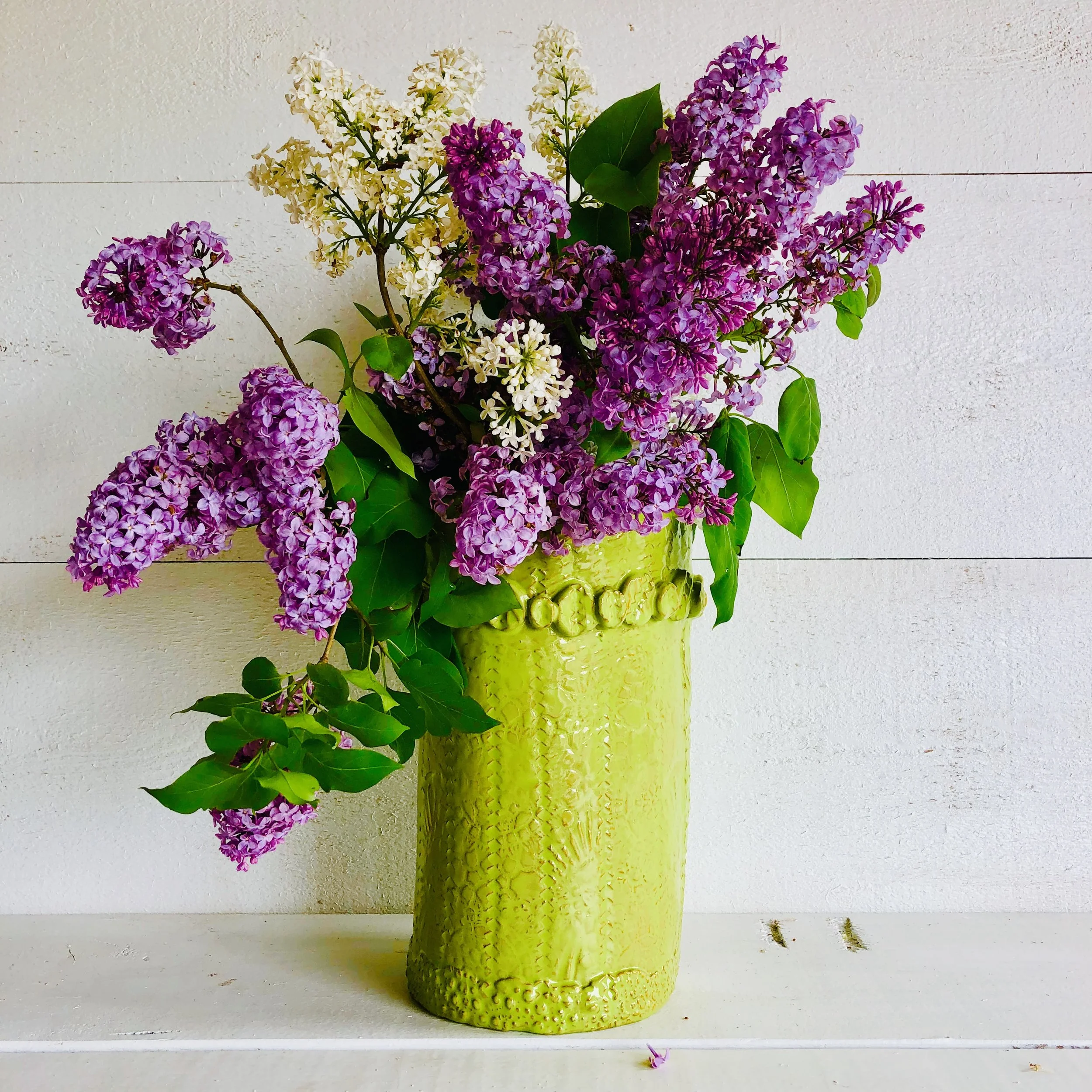 A bouquet of purple, white, and pink lilacs in a bright green textured ceramic vase placed on a white surface against a white wall background.