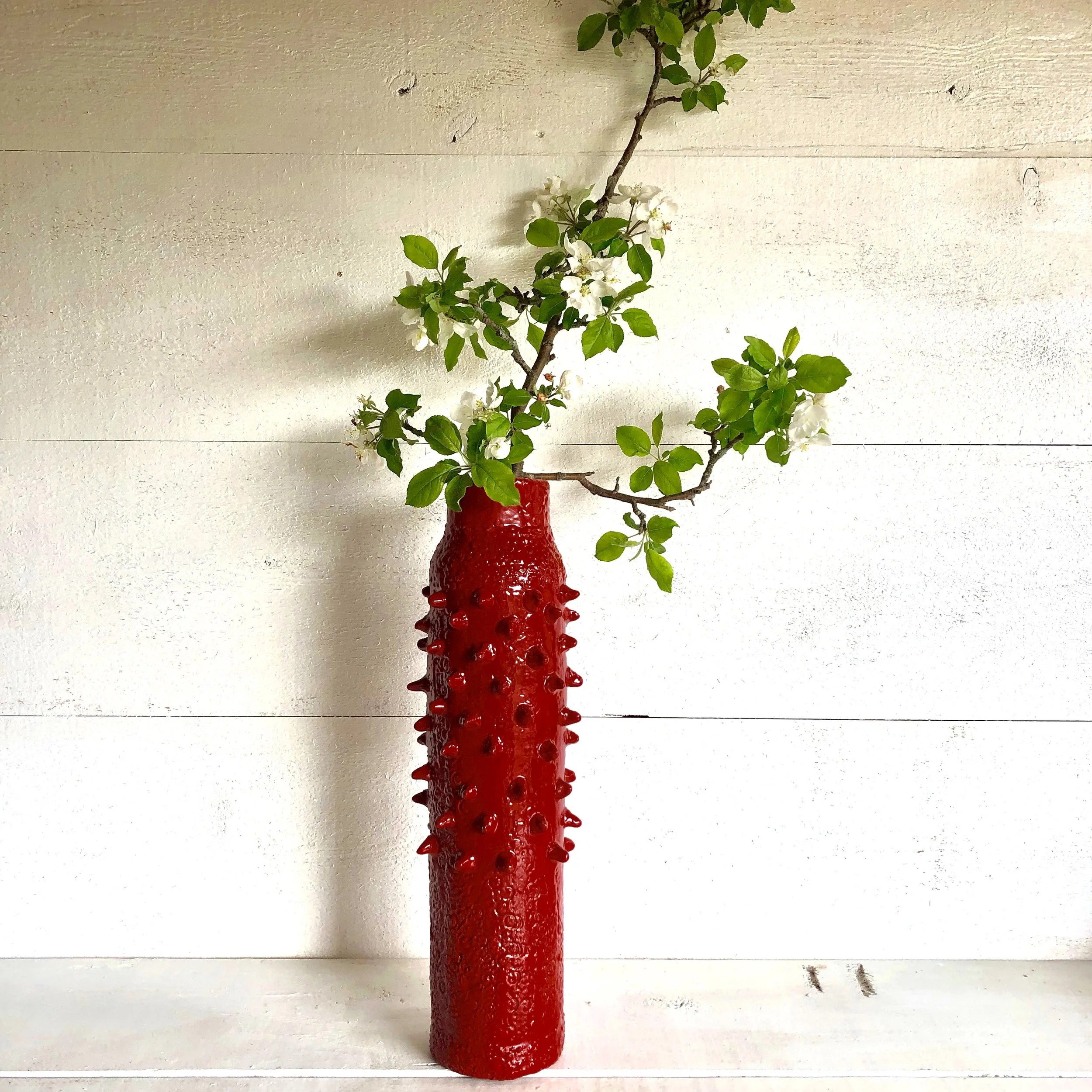 A tall, red, textured ceramic vase with small protrusions, holding a green leafy branch with white flowers, placed against a white wooden wall.