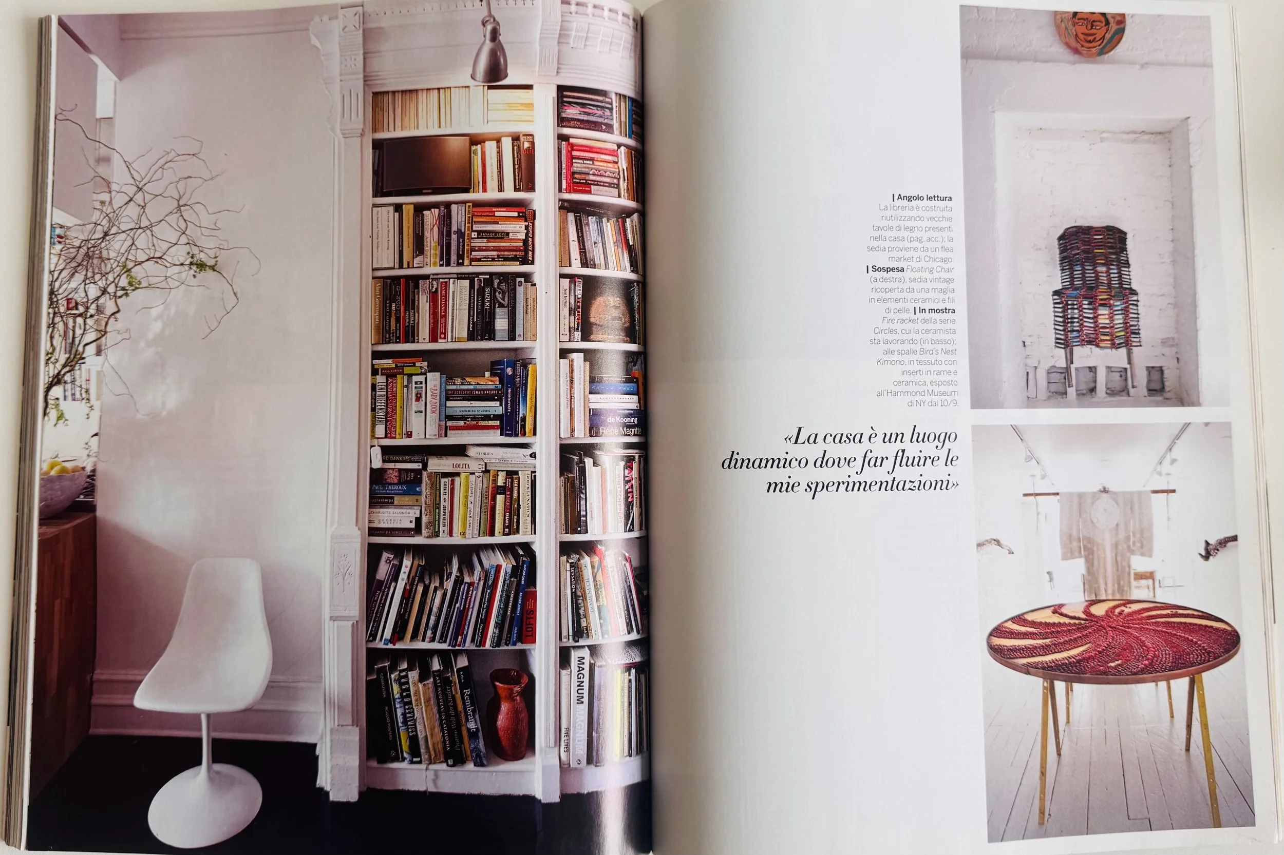 Open magazine showing interior decor with a white bookshelf filled with books, a white chair in front of the bookshelf, a branch with some leaves on the wall to the left, and two images on the right page: one of a colorful woven chair and another of a round table with a patterned top, a hanging textile, and ceiling lights.