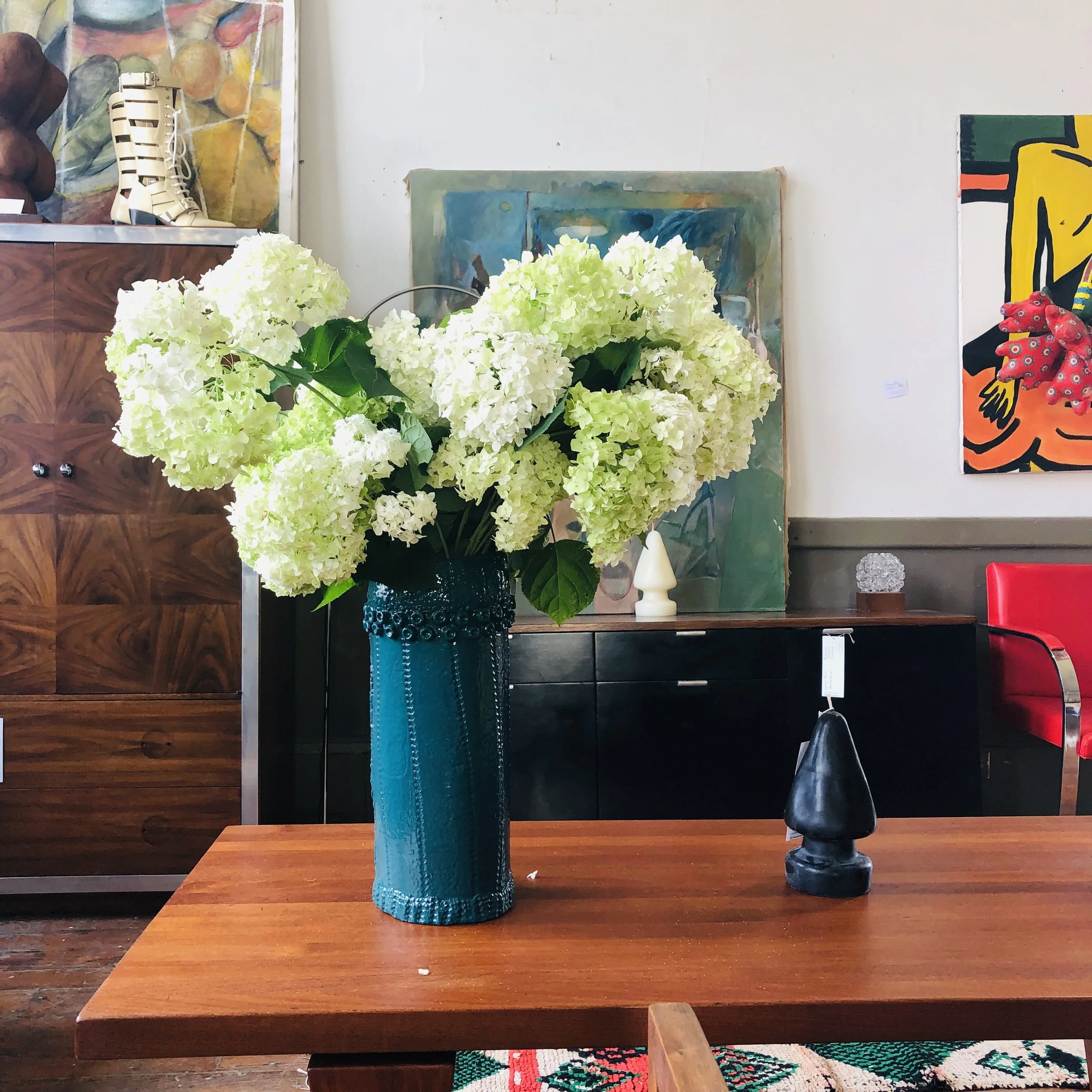 A tall teal vase filled with white and pale green hydrangea flowers placed on a wooden table in an art-filled room.