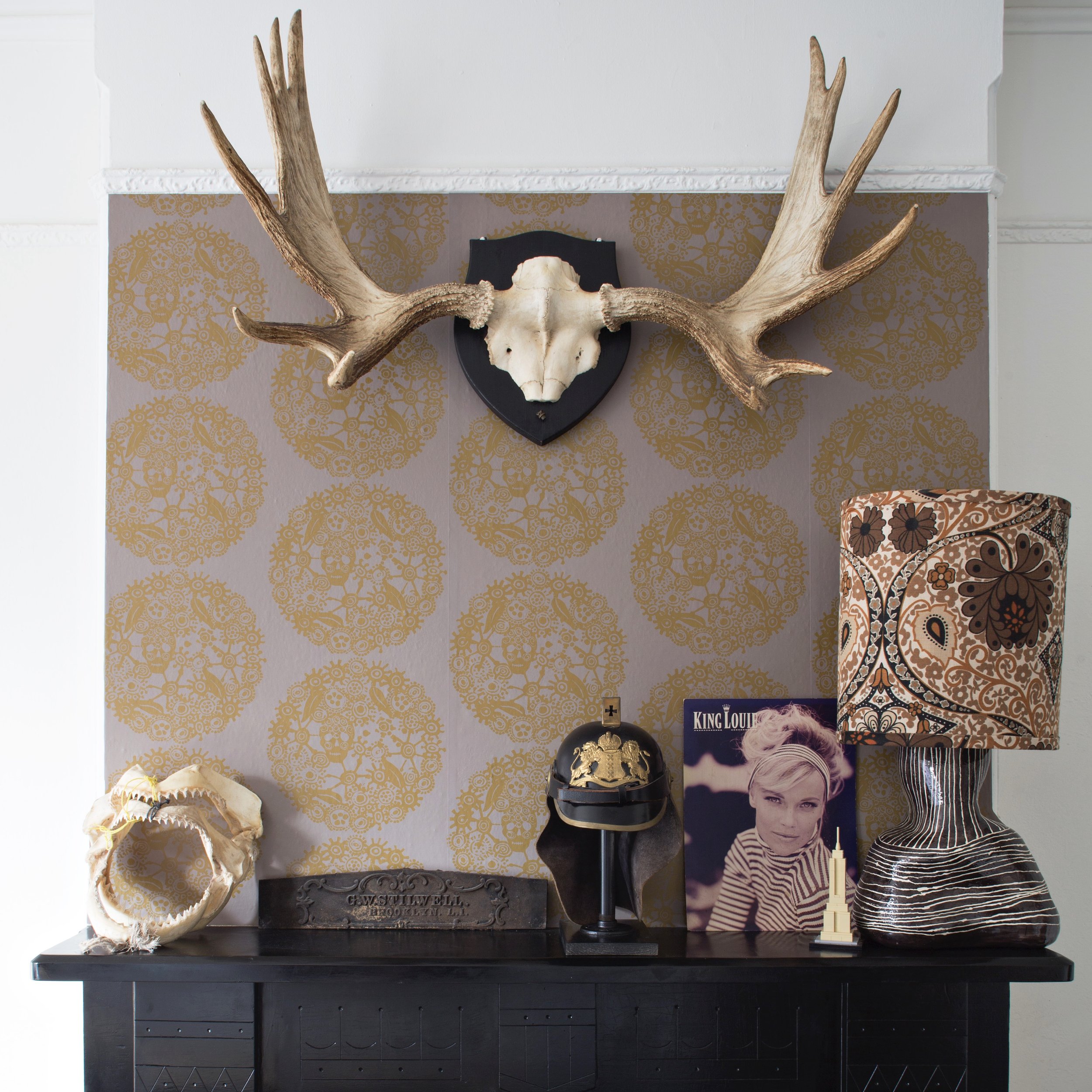 Decorative wall with animal skull and antlers, framed portrait of a woman, vintage helmet, ornate lamp, and other decor on black sideboard.