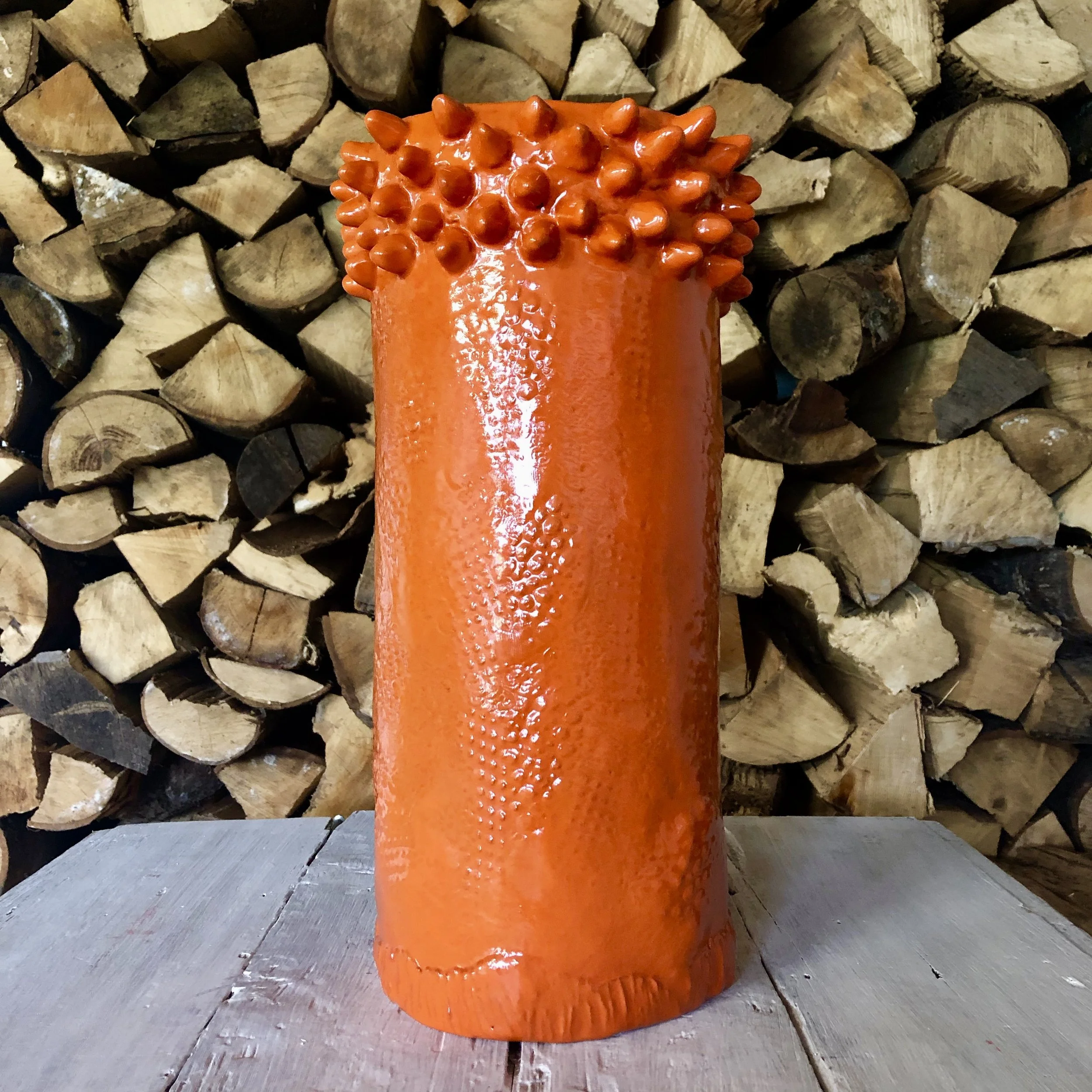 A tall orange ceramic vase with a textured surface and a cluster of small bulbous protrusions at the top, placed on a wooden surface with a background of stacked firewood.