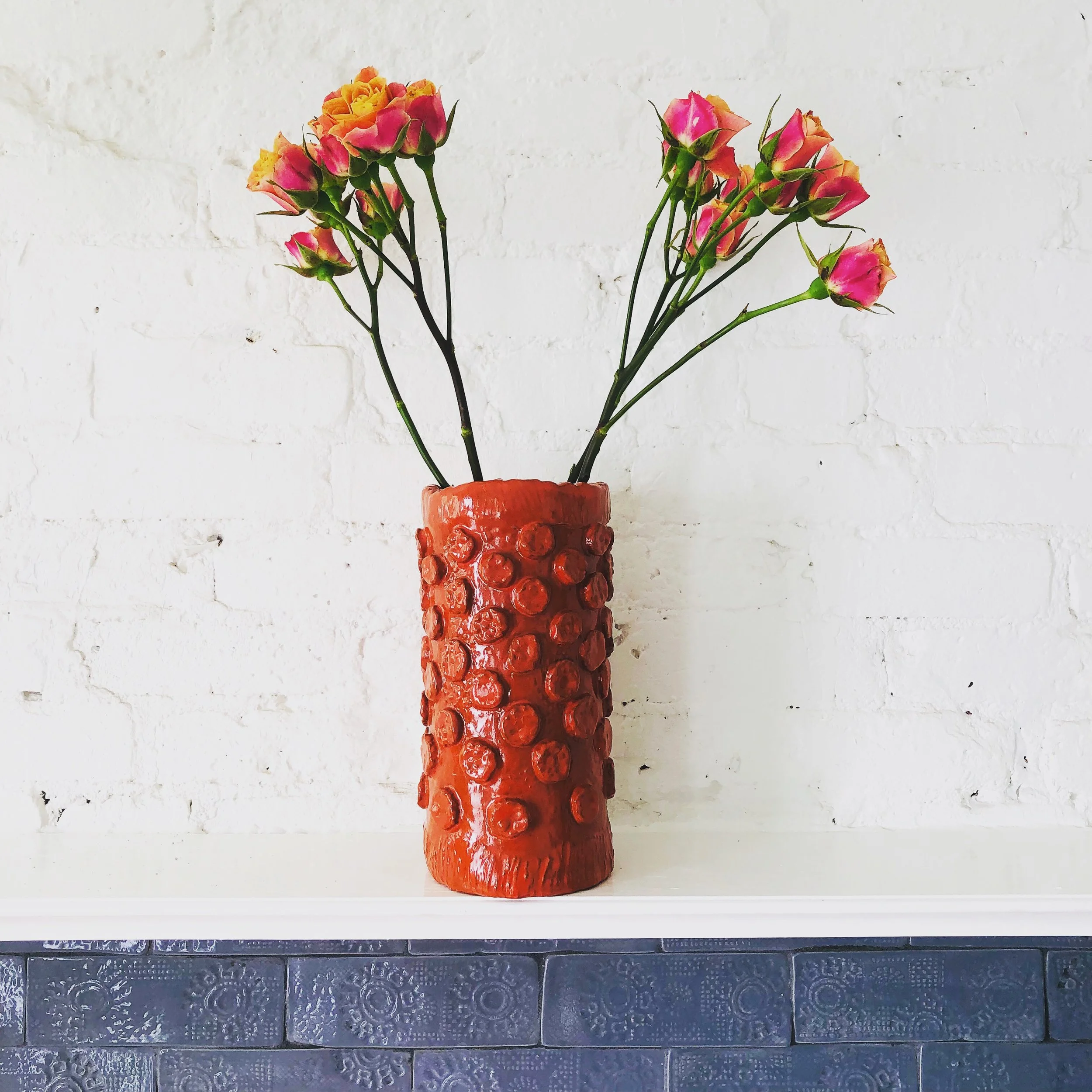A tall red ceramic vase with a textured, bubble-like surface, holding pink and yellow roses, placed on a white surface against a white brick wall.