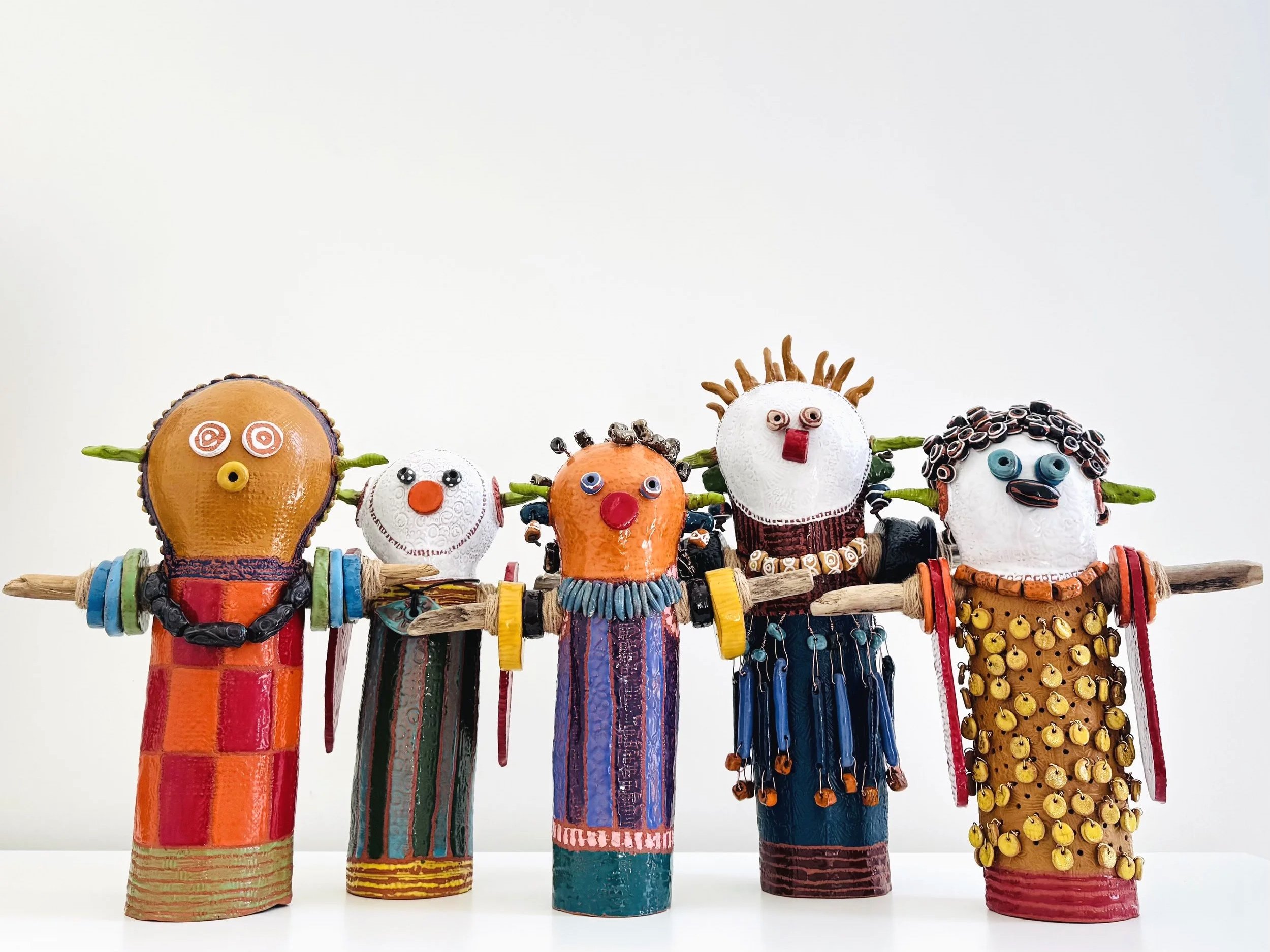 Colorful handmade figurines resembling humans with unique features, each holding a stick on their shoulders, decorated with various beads, shells, and materials.
